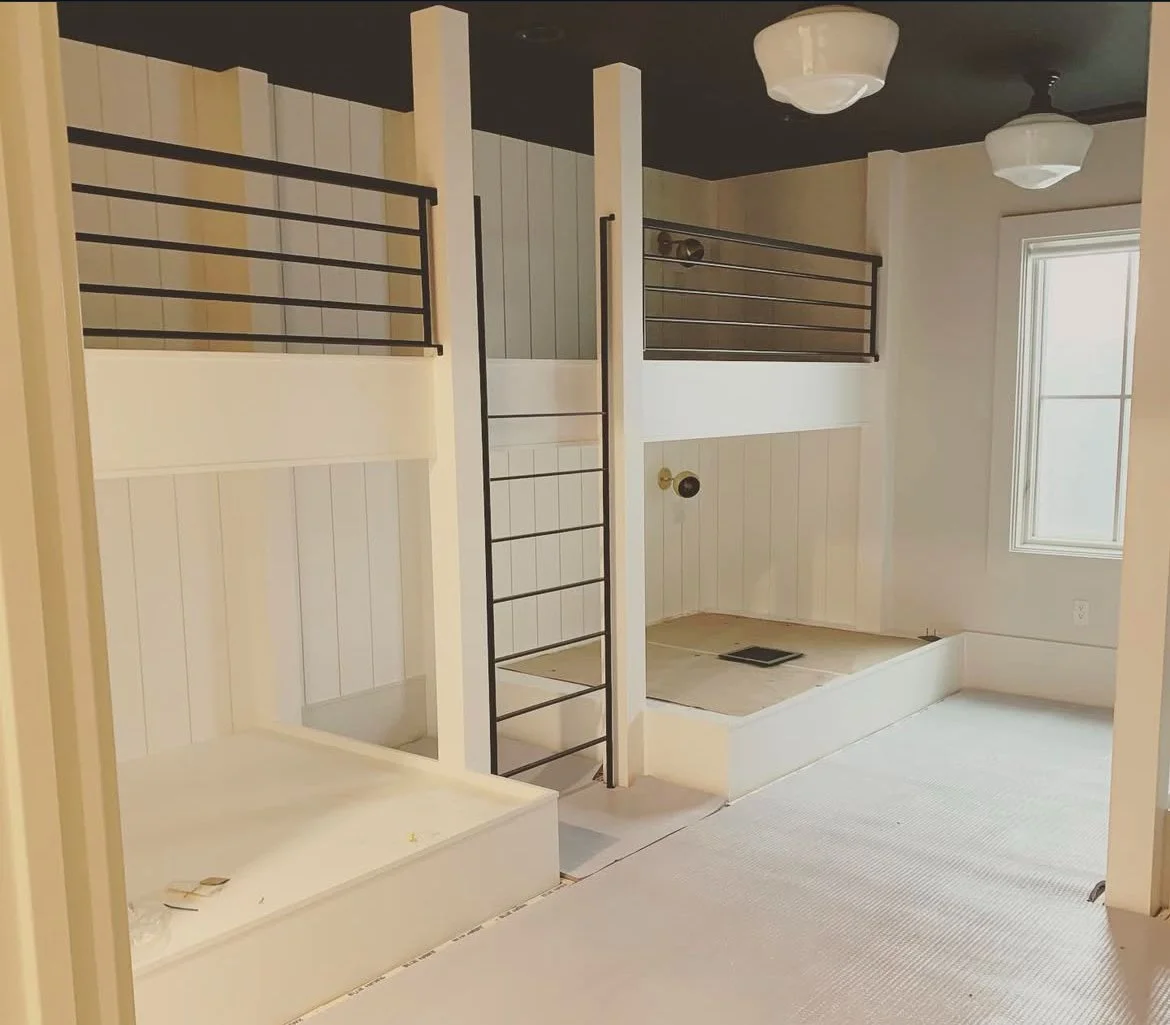 A room with two small raised platform areas, one with a ladder, and black metal railings, with beige walls, a window, and ceiling lights.