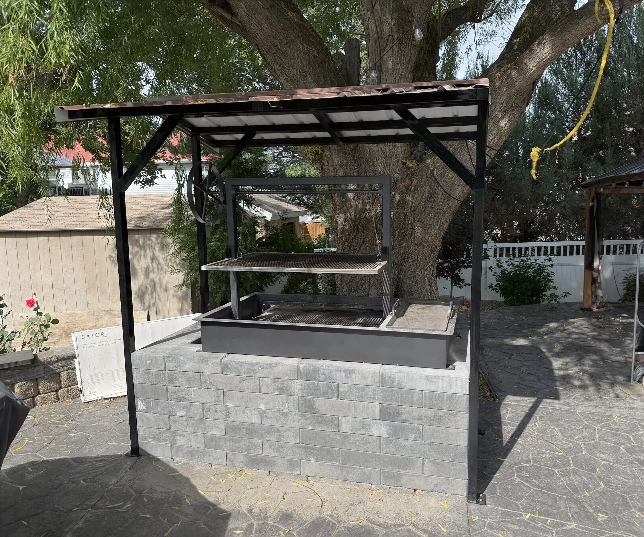A modern outdoor brick fireplace with a metal cooking grill in a backyard, surrounded by trees and a white fence.