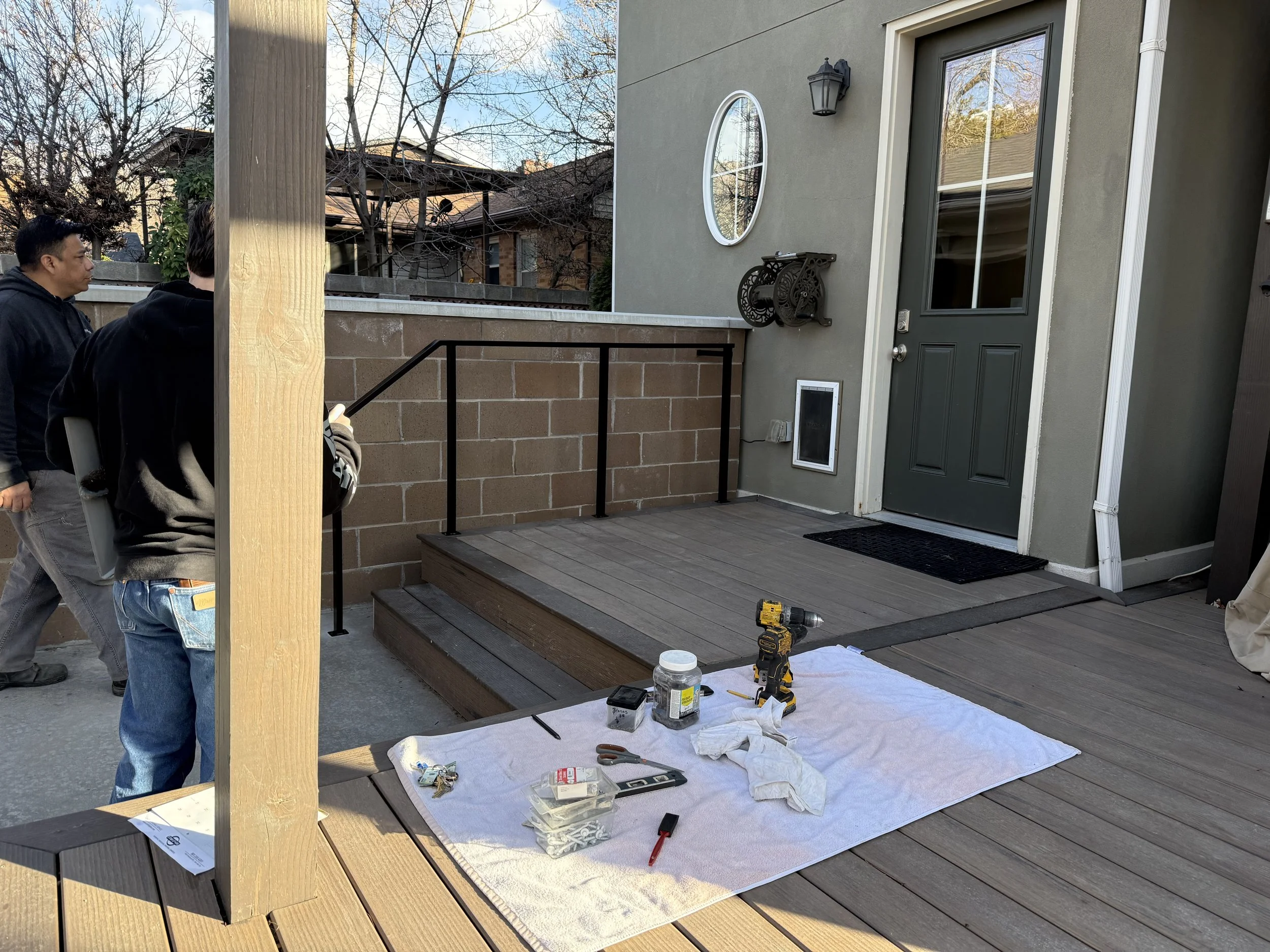 People working on a small wooden deck next to a house with green door and exterior wall, tools and supplies laid out on a white cloth, construction materials, and a ladder nearby.