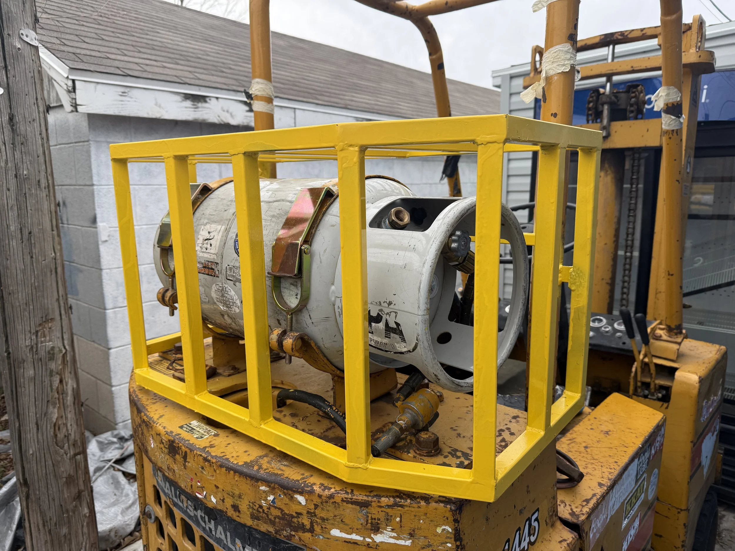 Industrial lift equipment with a yellow safety cage, engine, and mechanical parts, mounted on a yellow platform, outdoors near a building with a wooden post and siding.