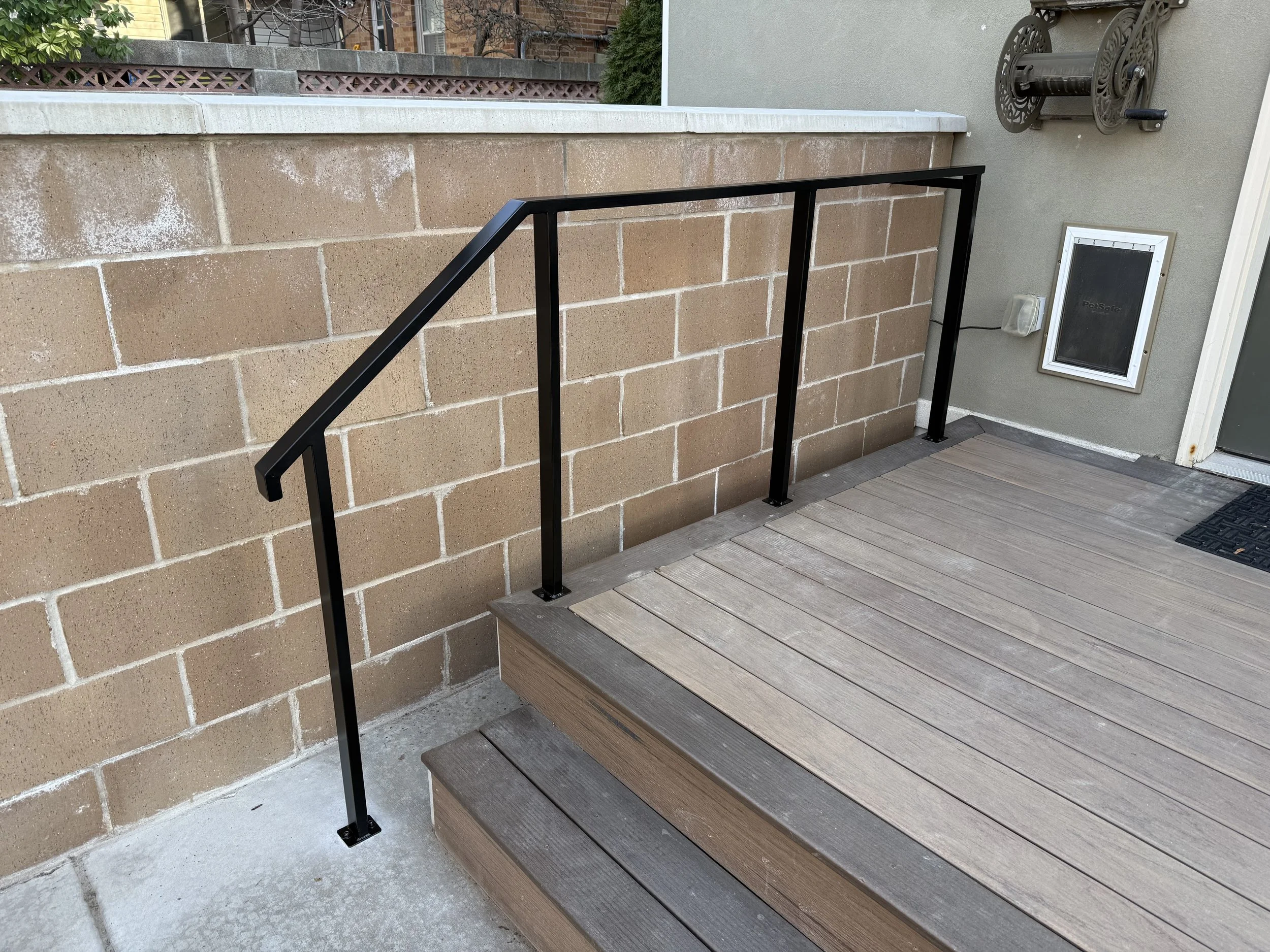 Image of an outdoor deck area with stairs, a brick wall with brownish-beige blocks, a black metal railing, a concrete floor, a light gray wall with a PetSafe pet door, and a hose reel attached to the wall.