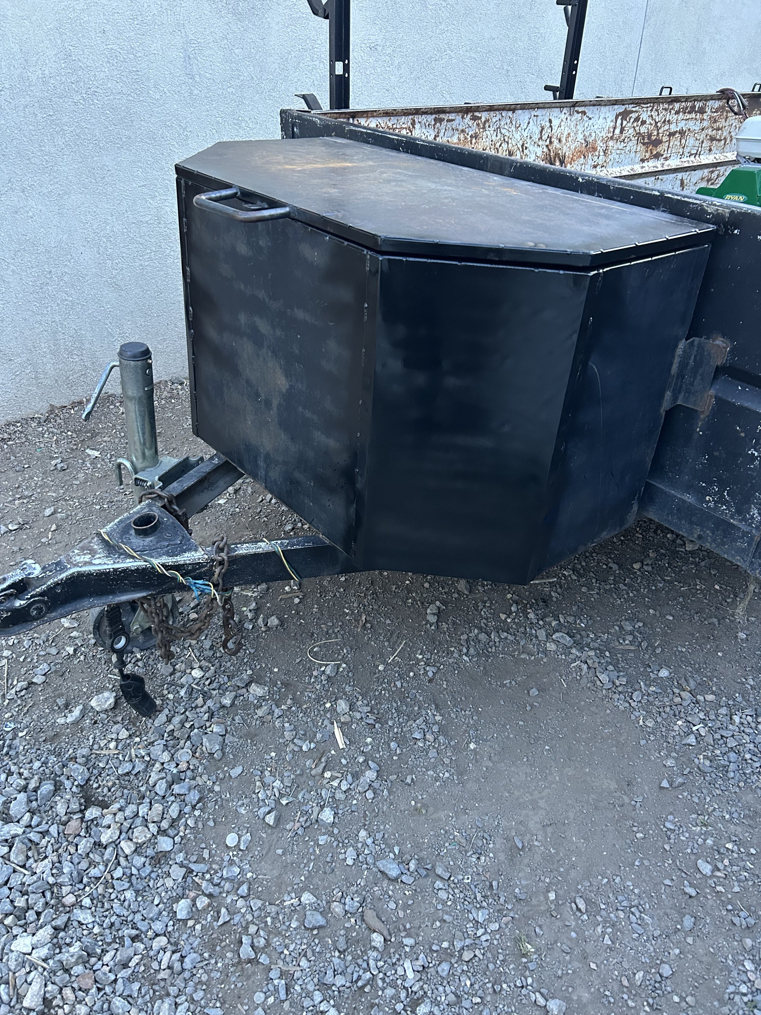 A black utility box mounted on a trailer hitch, with a rusty metal container in the background, on gravel ground next to a light-colored wall.