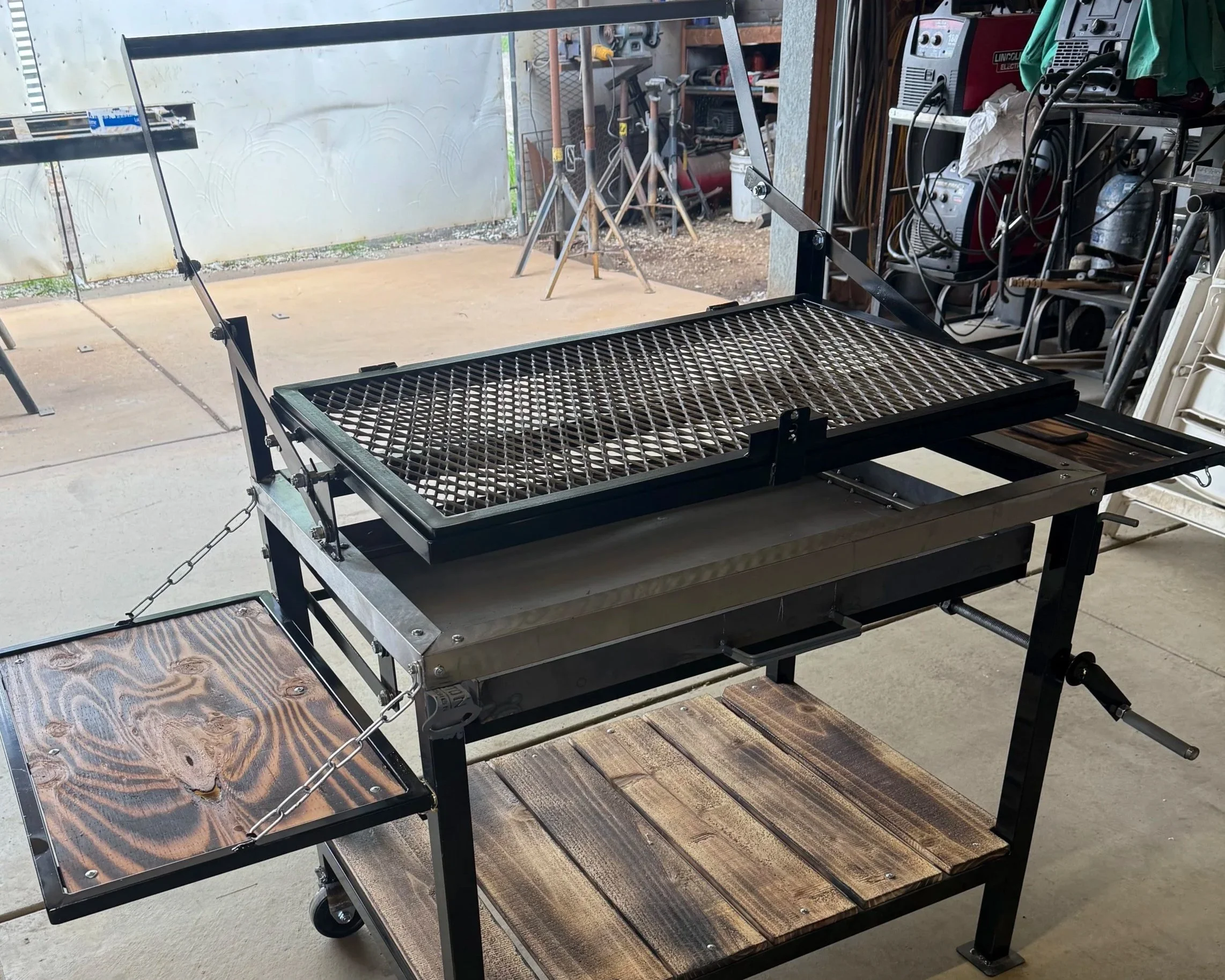 A black metal grill or smoker on a wooden table in a garage or workshop setting, with a metal exhaust hood and various tools and equipment in the background.