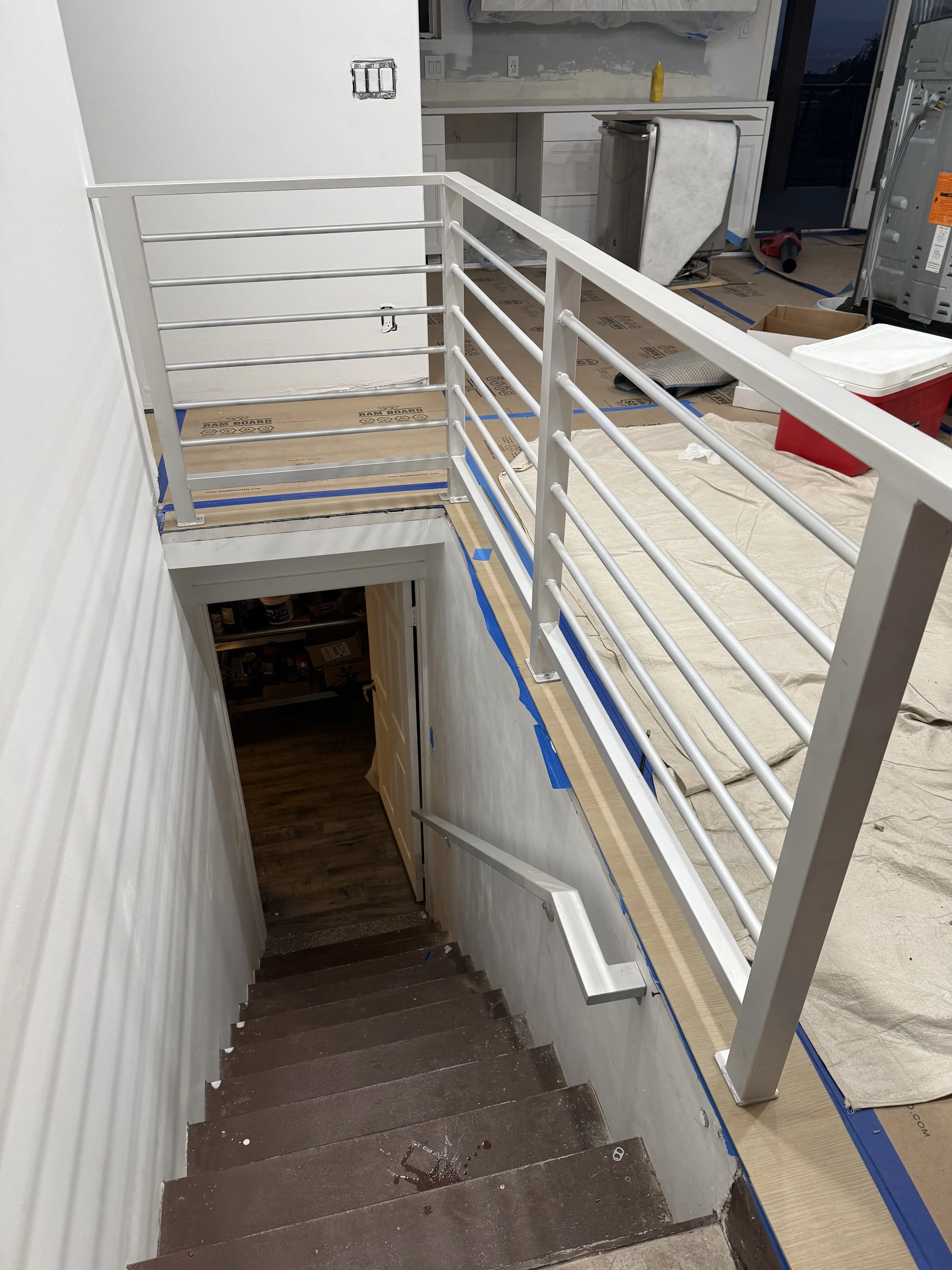 Interior staircase with a new white metal safety railing, seen from the top looking down. The stairs are brown, leading to a lower level, with some construction or renovation materials visible around, including a partially covered countertop and a red cooler.