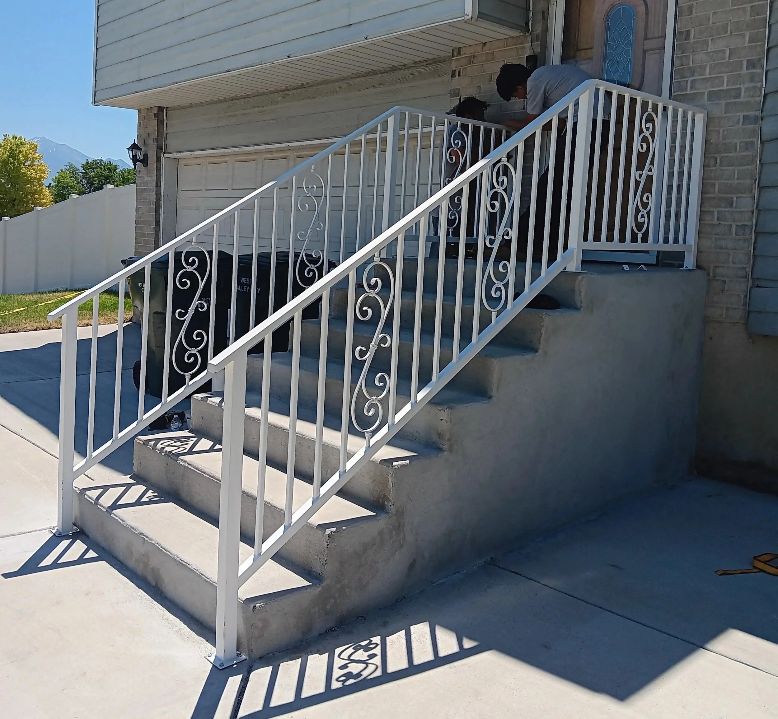 Two people working together on a small porch, with one leaning over and the other looking on. White metal railing and gray brick exterior wall of a house.