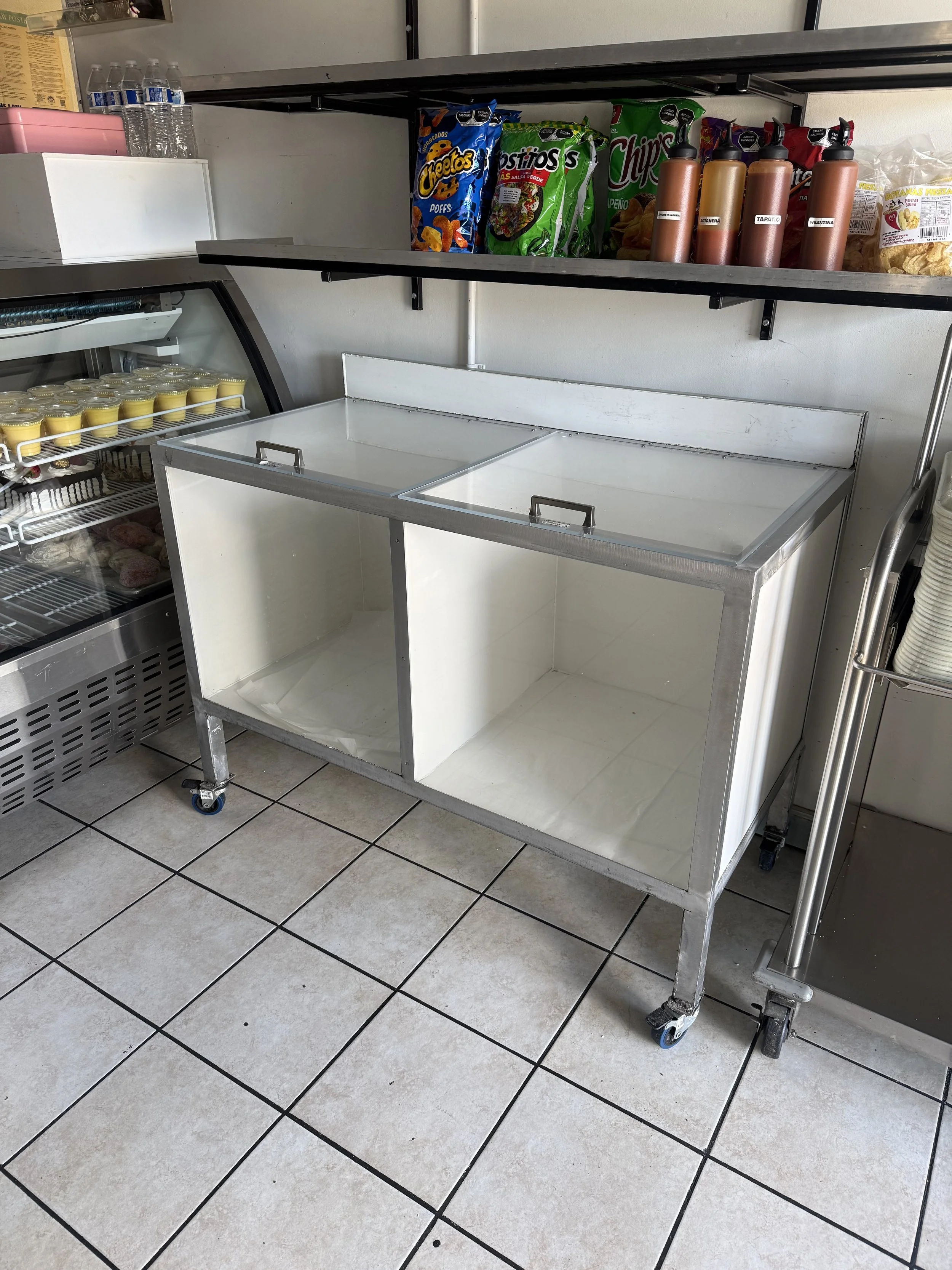 Empty commercial display cooler with open sliding glass doors, positioned in a tiled store or restaurant setting. Shelves above hold bottled water, snack bags, and bottles of salad dressing. The cooler has a metal frame with wheels at the bottom.