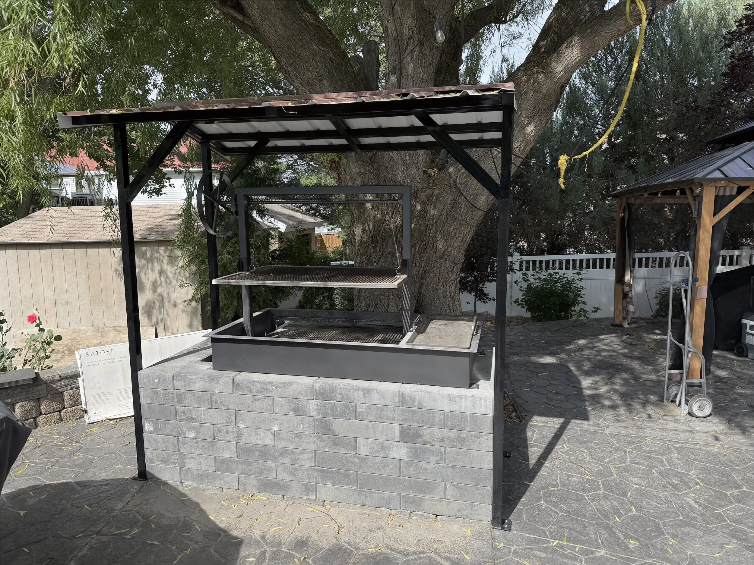 An outdoor garden area with a granite counter, a swing, and a metal-framed barbecue grill underneath a roofed shelter.