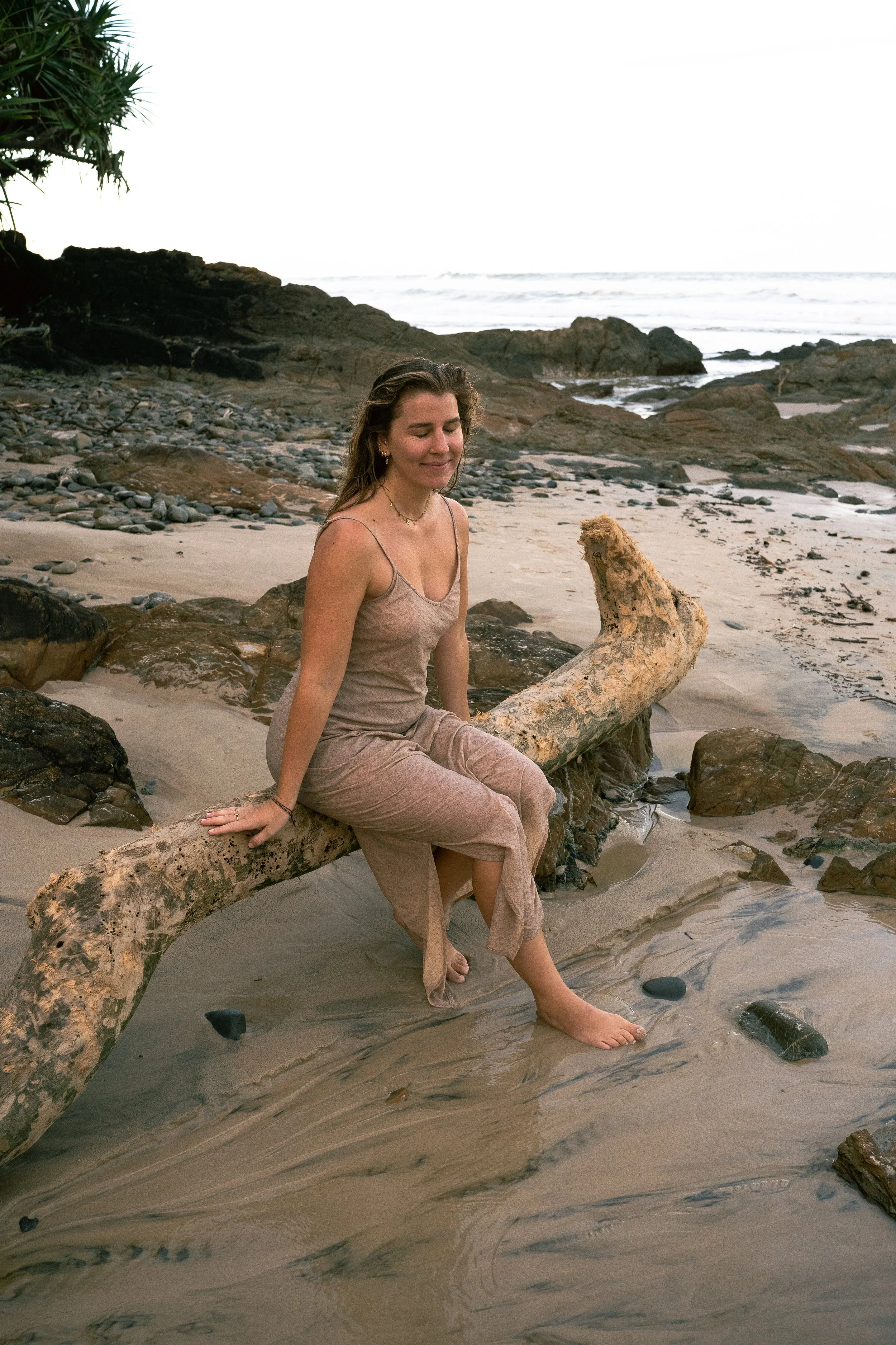 A woman sitting on a large driftwood log on a sandy beach, with rocks and ocean waves in the background.