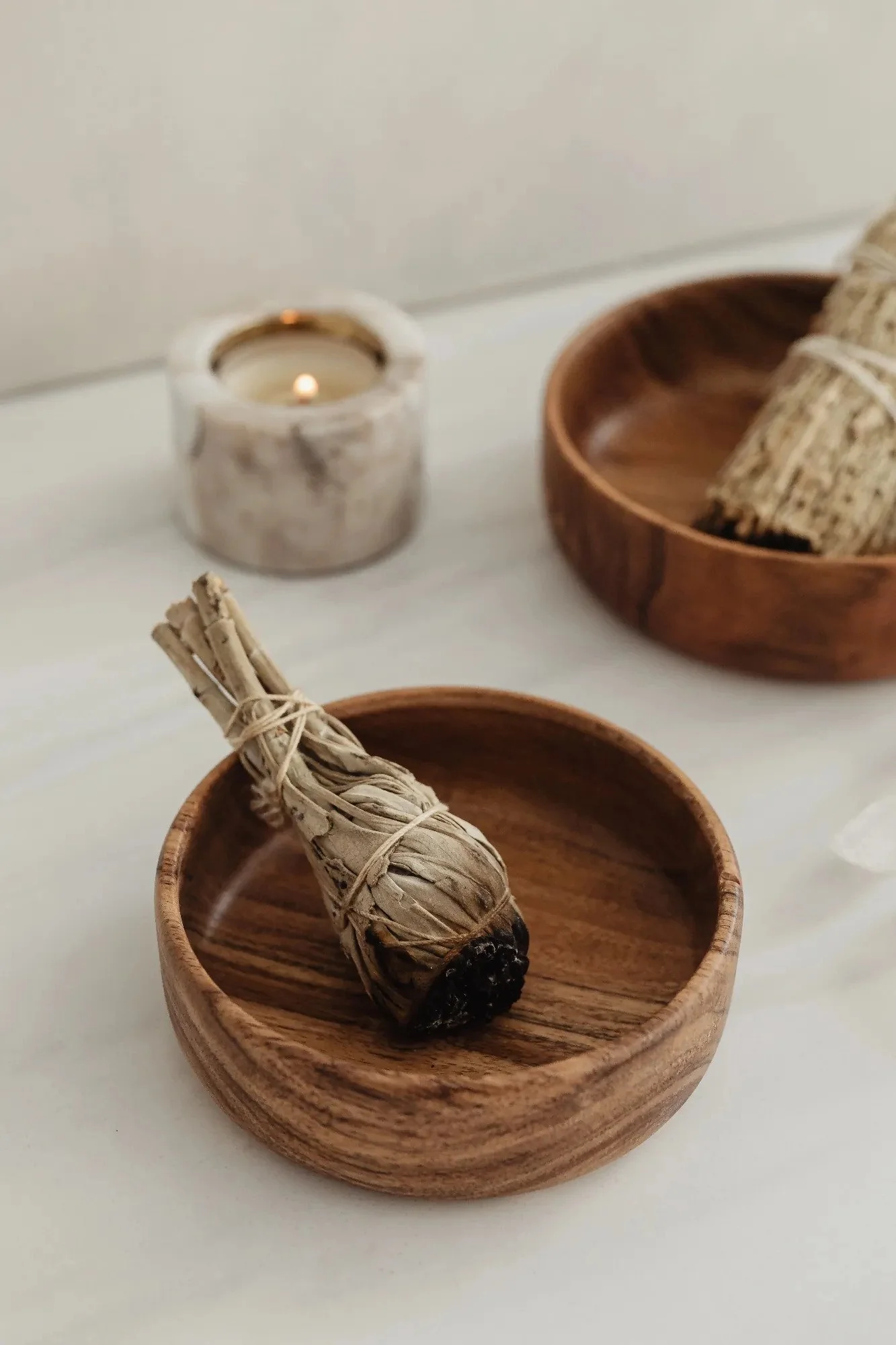 A smudge stick wrapped in twine in a wooden bowl, with a lit candle and other smudge sticks in wooden bowls in the background.