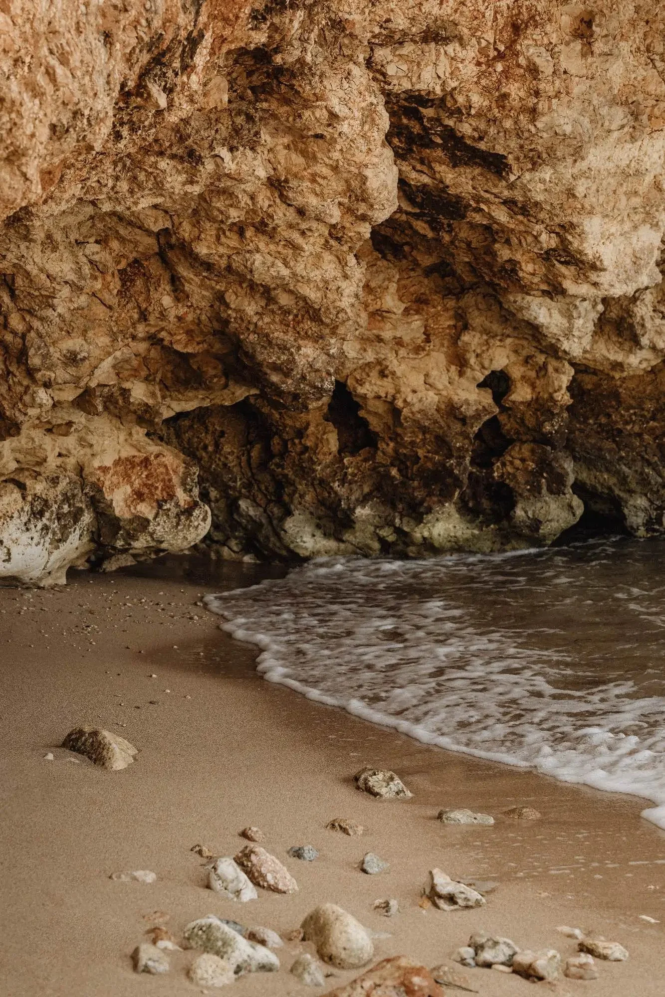 A sandy beach with small rocks in the foreground, and a large rocky cliff in the background with water gently lapping at the shore.