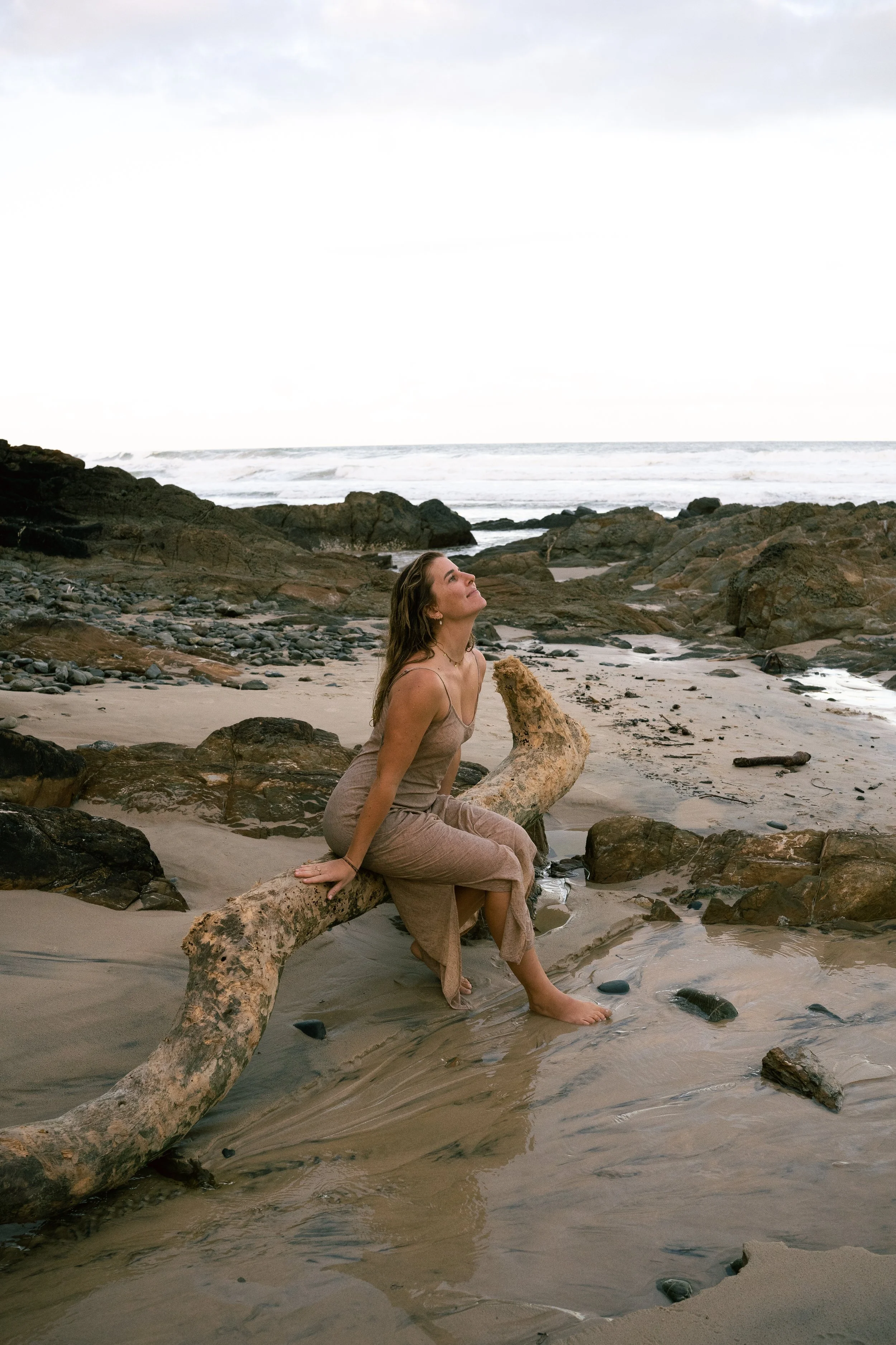 Woman sitting on a log on a rocky beach, looking up at the sky.