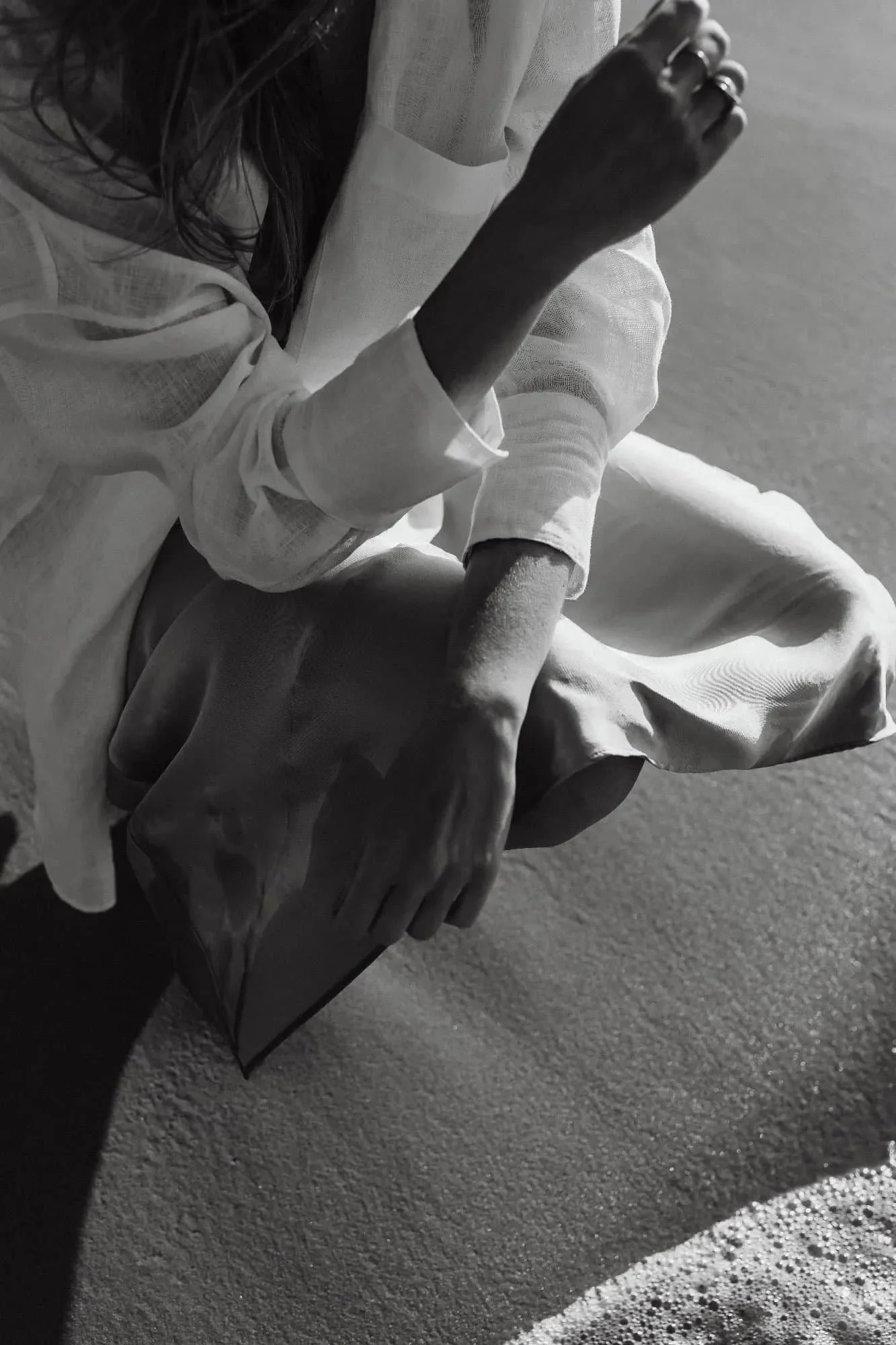 Black and white photo of a person sitting on a beach, holding a rectangular object, with waves in the foreground.