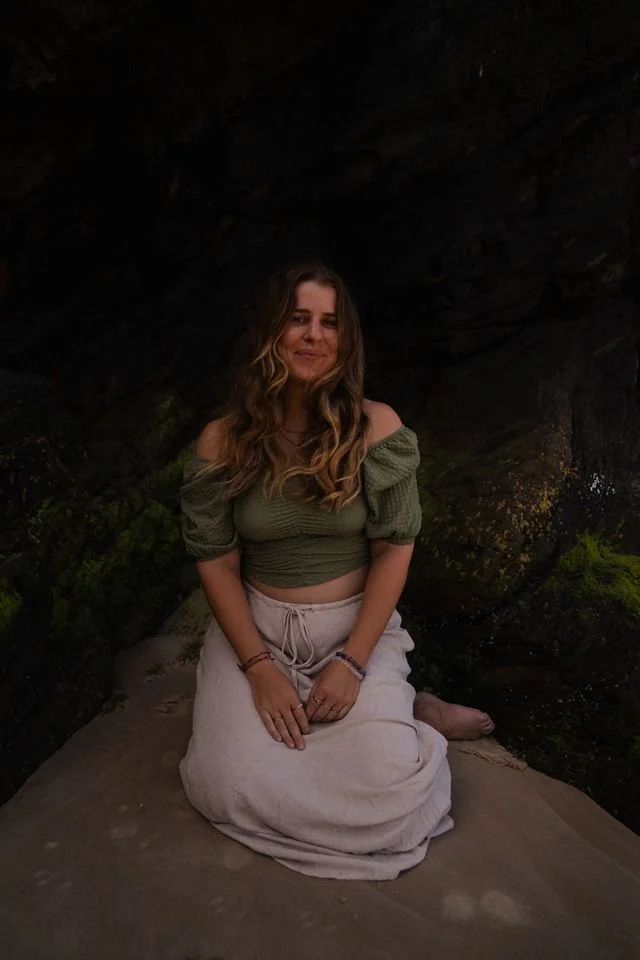 A woman kneeling on a sandy surface in front of dark rock formations, wearing a green off-shoulder top and light-colored skirt, smiling softly.