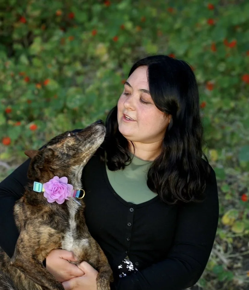 A woman with black hair holding a brindle dog with a pink collar and a flower decoration, both looking affectionately at each other outdoors.