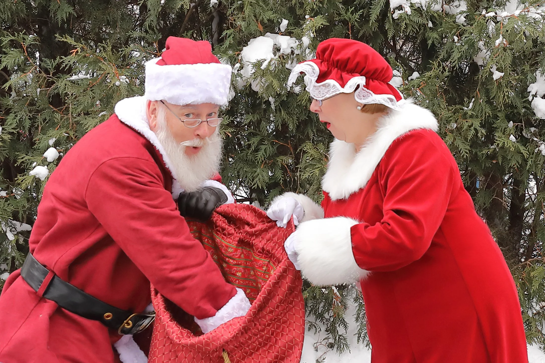 Santa Claus and a woman in Christmas costumes exchanging a gift outdoors with a snow-covered evergreen tree in the background.