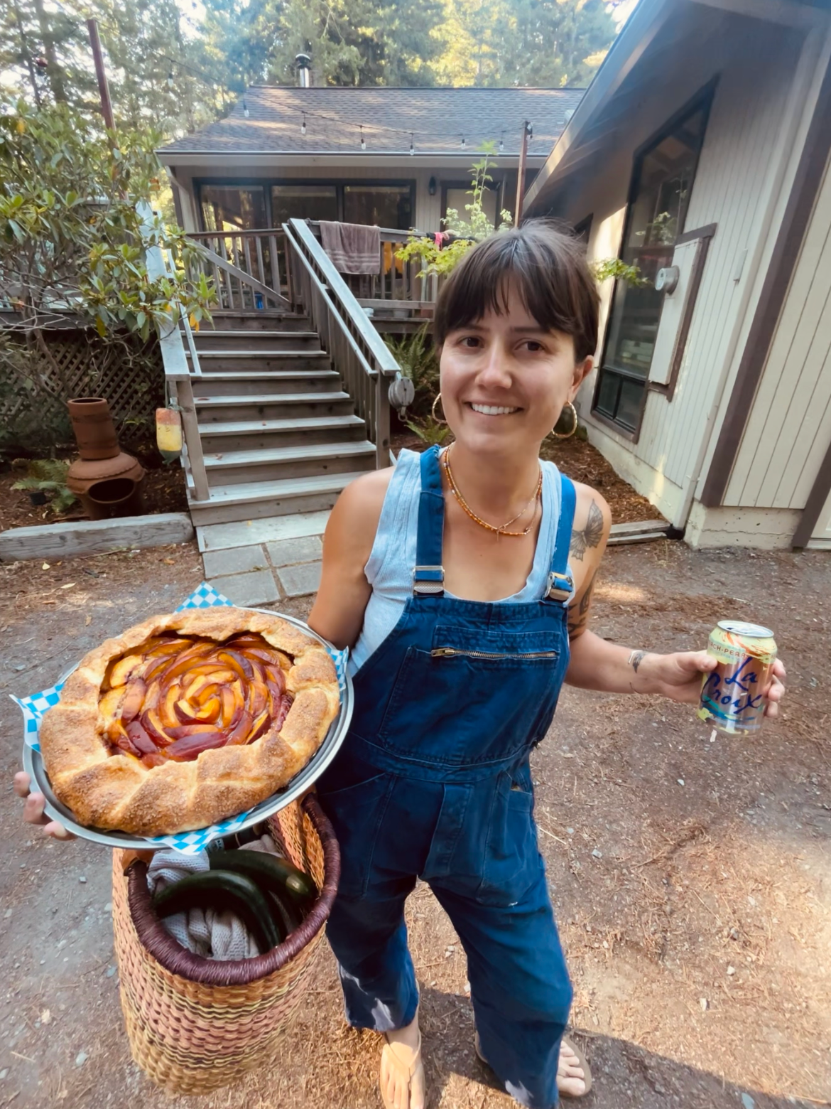 A woman stands outdoors holding a plate with a large peach-cobbler-like pie and a can of LaCroix sparkling water, smiling at the camera.