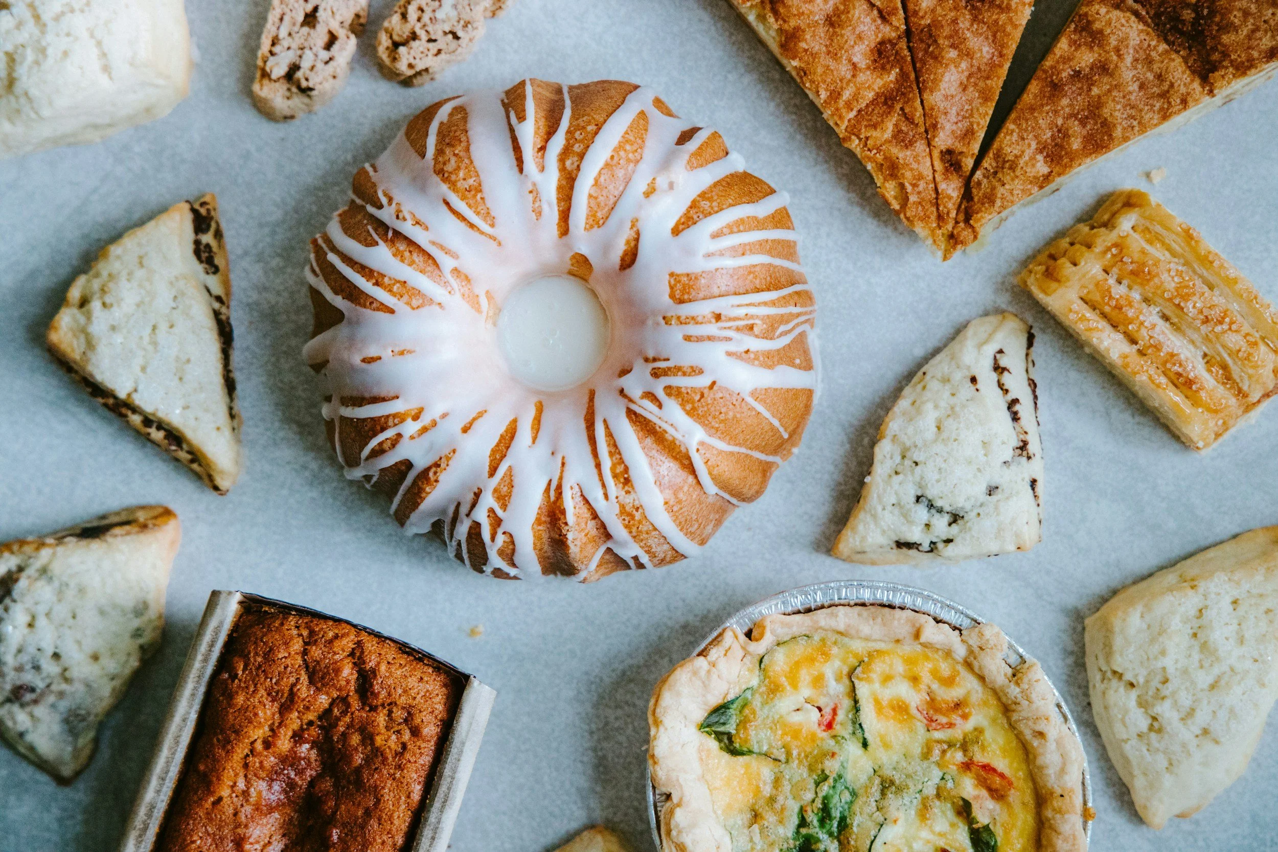 Assorted baked goods including a glazed donut with white icing, slices of cake, a quiche, and various cookies on parchment paper.