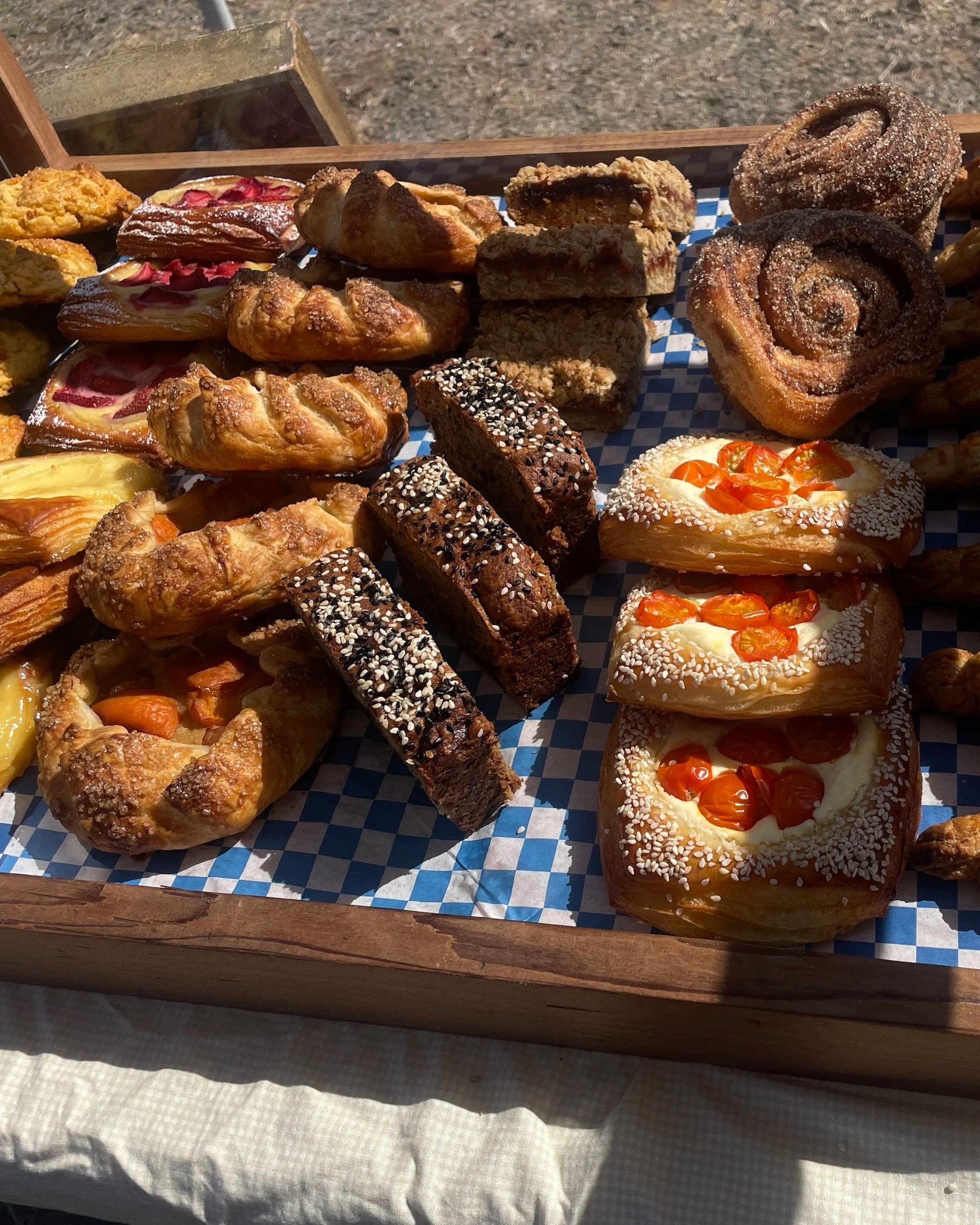 Assorted baked goods including cinnamon rolls, brownies with white sprinkles, fruit Danish pastries, cinnamon twists, and bars with fruit toppings on a blue checkered paper.