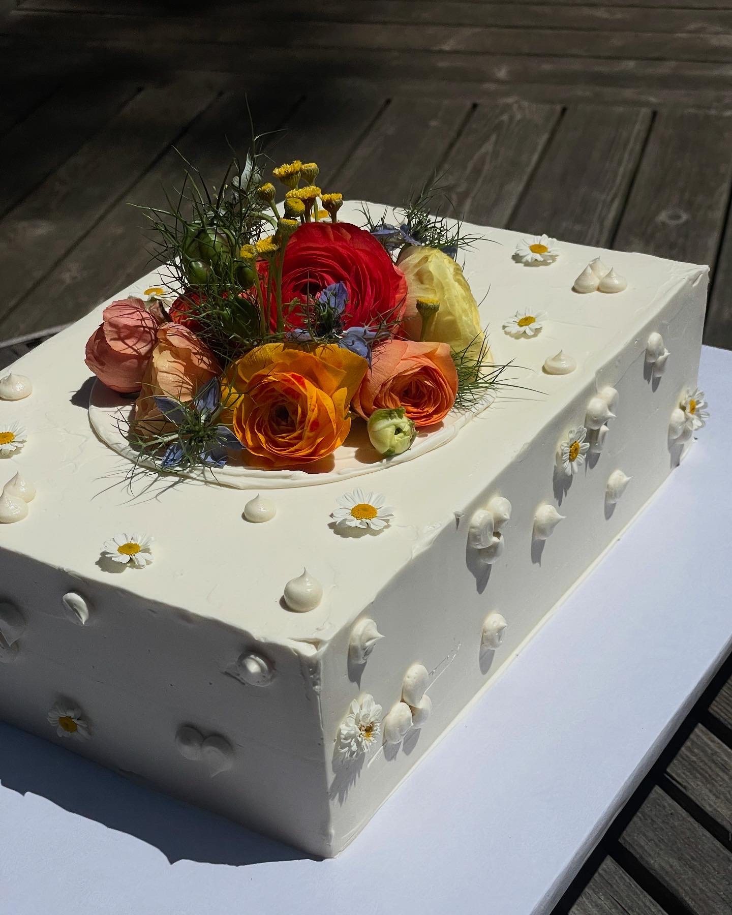 Rectangular white cake decorated with white icing, small daisy-like flowers, and topped with various colorful fresh flowers.