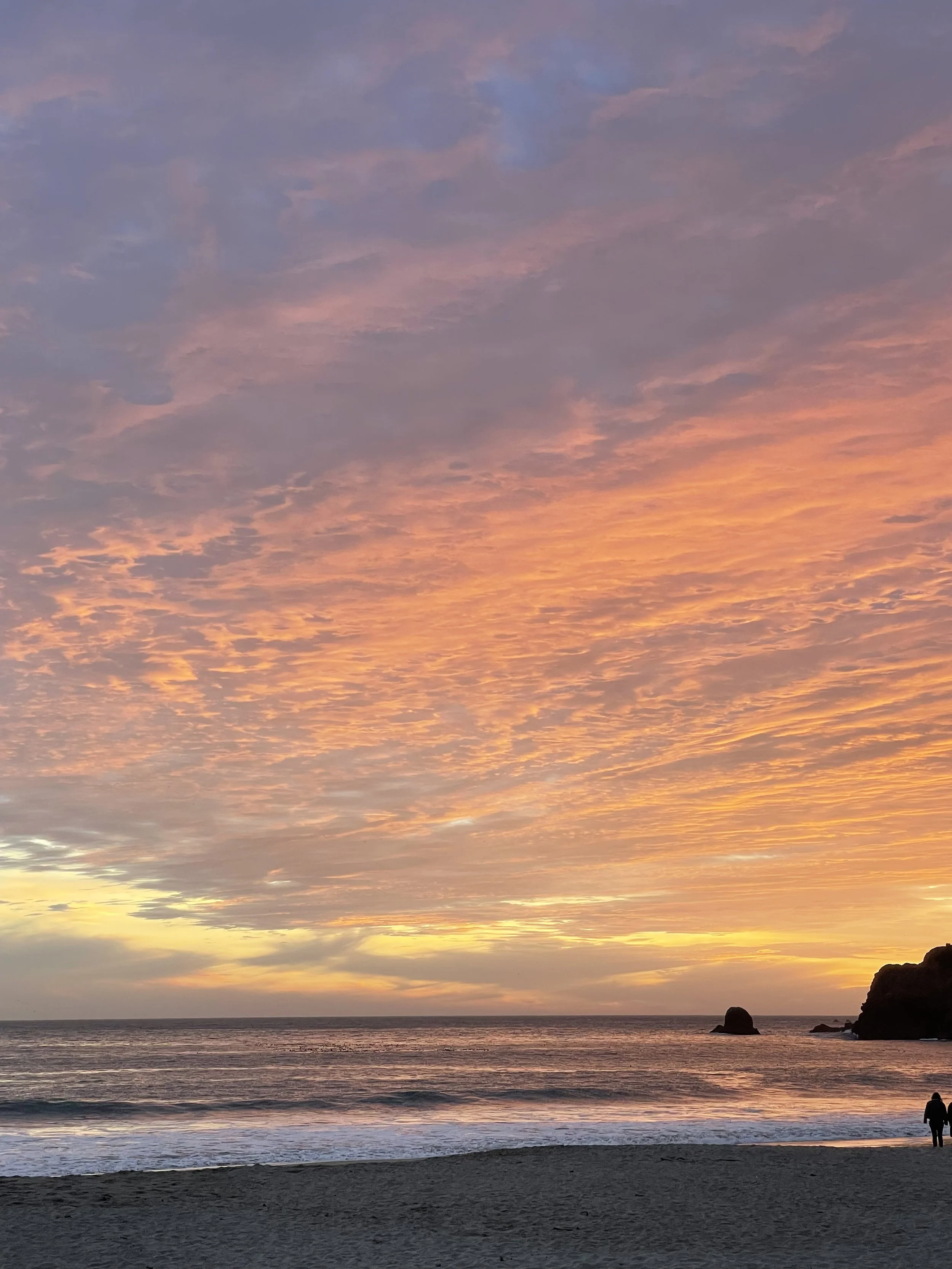 Sunset over a beach with orange and purple clouds, dark ocean waves, and two people walking along the shore.