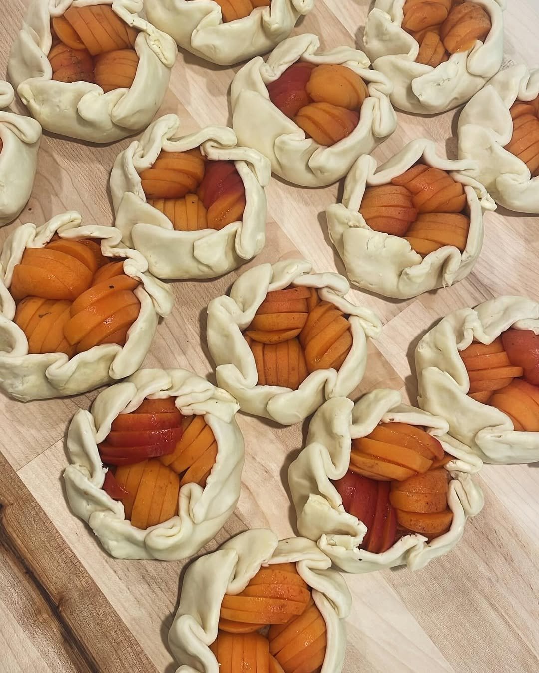 Multiple peach and purple stone fruit tarts with crimped pastry crusts laid out on a wooden surface.