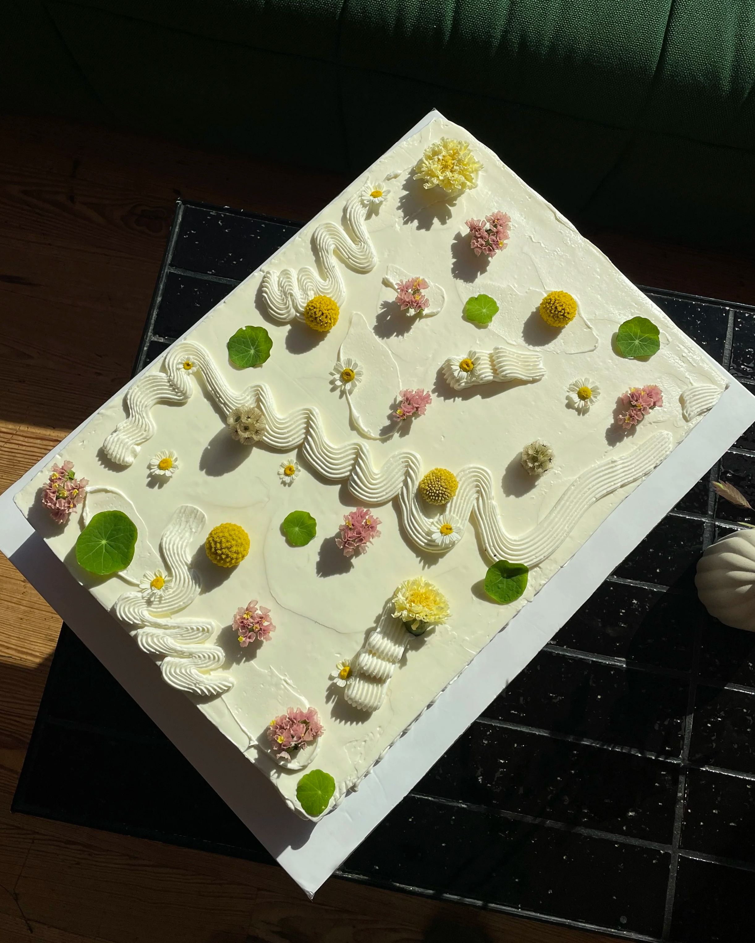 Rectangular white cake decorated with various colorful flowers and green leaves, with a swan-shaped outline in the center, on a black tiled table.
