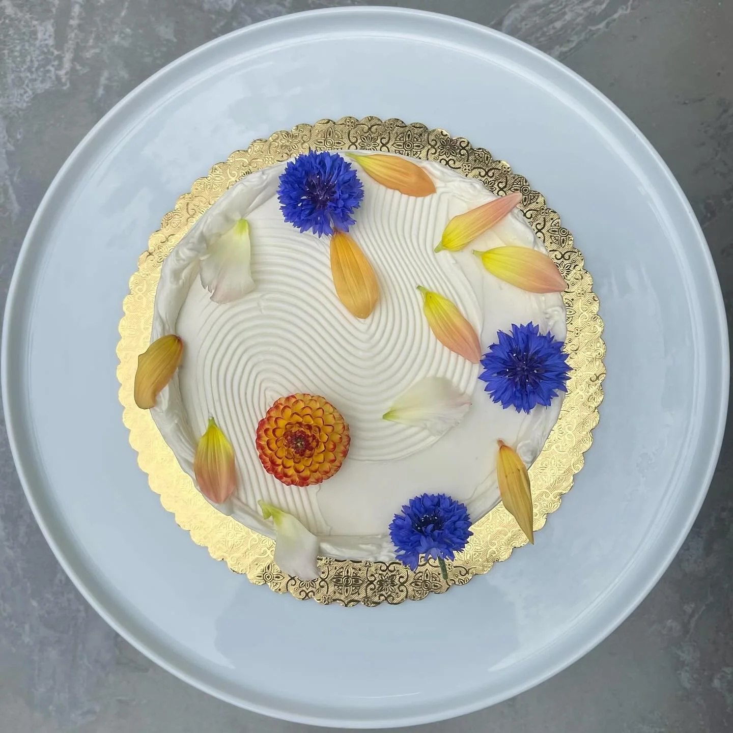A round white frosted cake decorated with blue, pink, yellow flowers and petals, placed on a gold decorative base on a white tray.