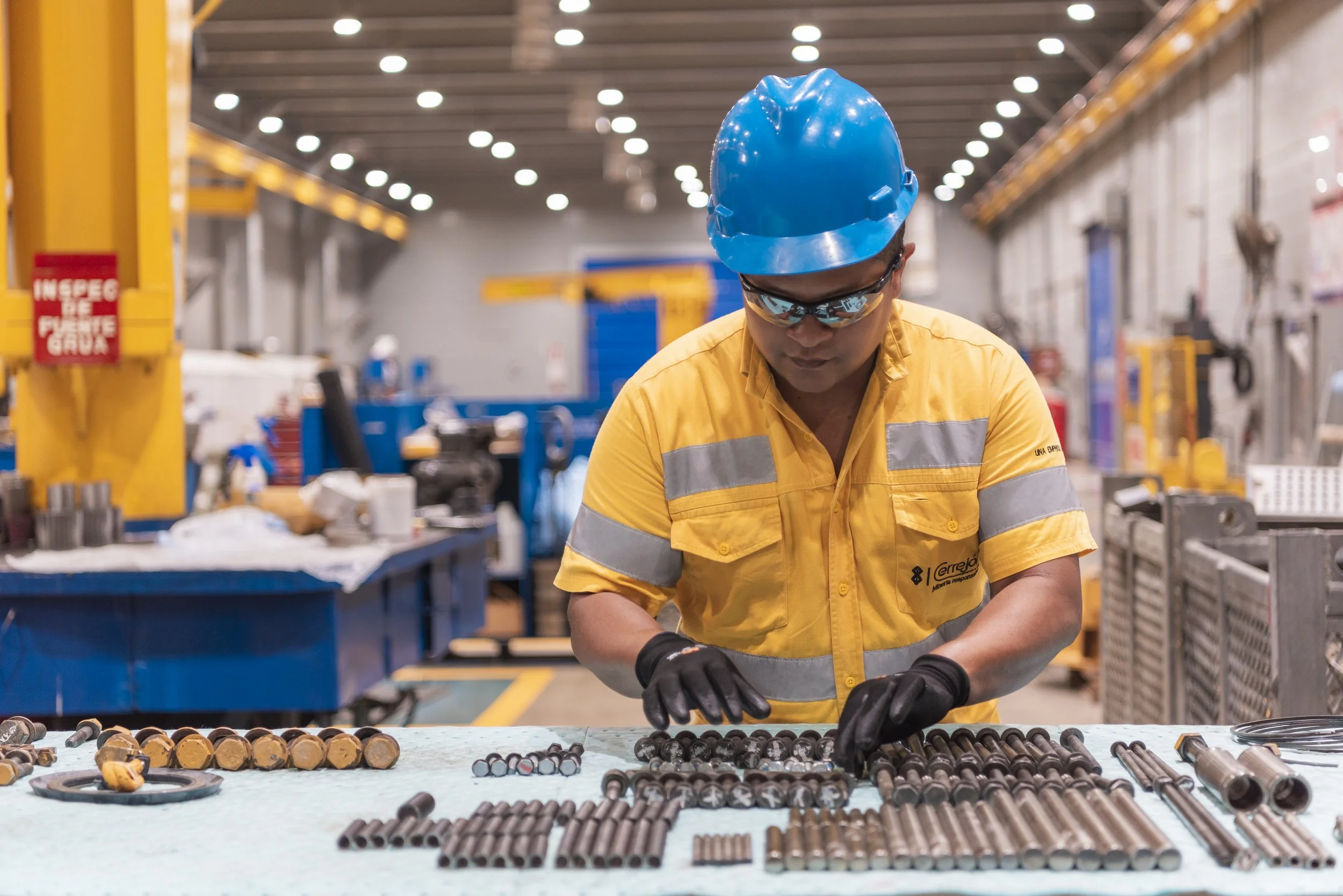 Un trabajador en equipo de protección incluyendo casco, lentes y guantes, inspecciona componentes metálicos en una línea de producción en una fábrica industrial.
