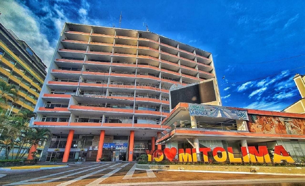 Edificio moderno de varios pisos con cartel que dice 'Yo ❤️ Tolima' en la entrada, cielo azul y nubes en el fondo.