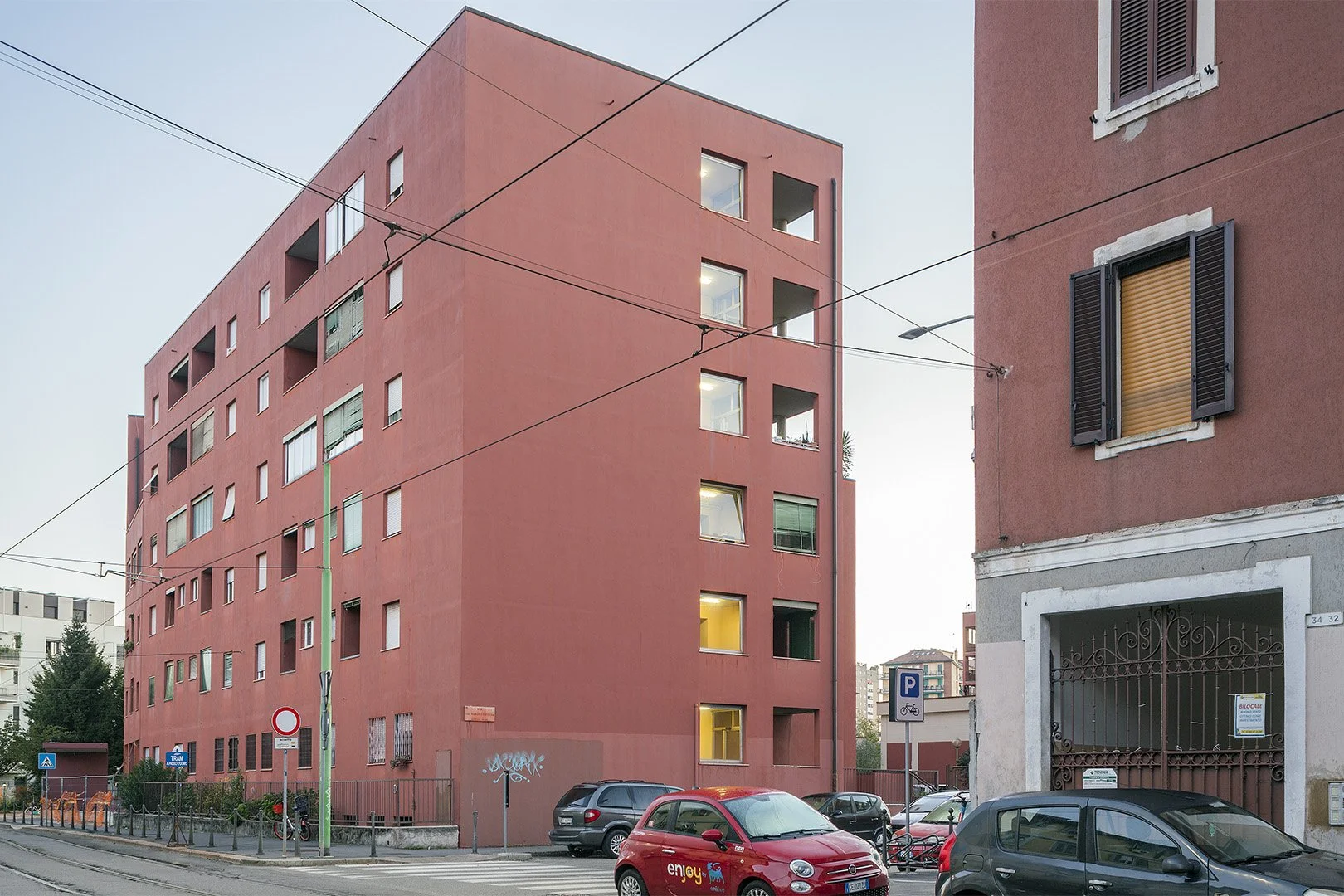 Edificio residenziale di colore rosa con alcune finestre illuminate, veicoli parcheggiati davanti e cartelli stradali, tra cui uno di divieto di sosta.
