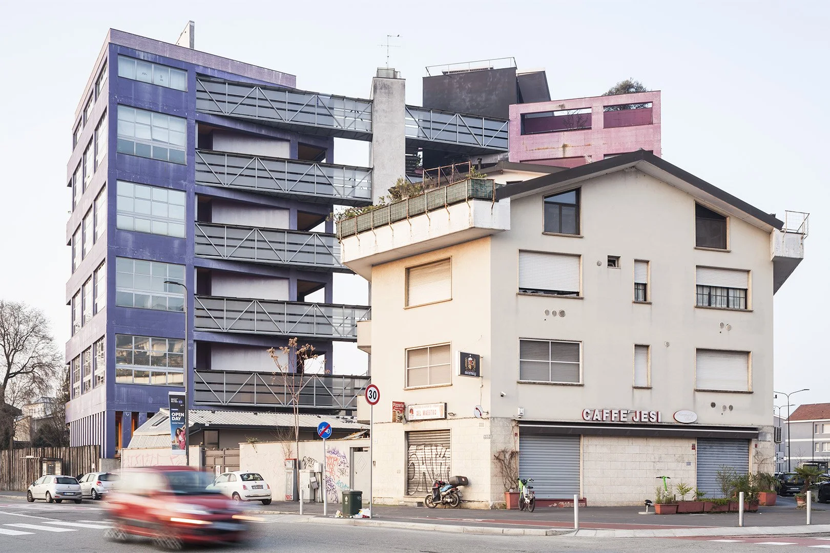 Due edifici urbani, uno moderno e uno vecchio, affacciati su una strada con auto in movimento, pneumatici e biciclette parcheggiate, sfondo con cielo chiaro.