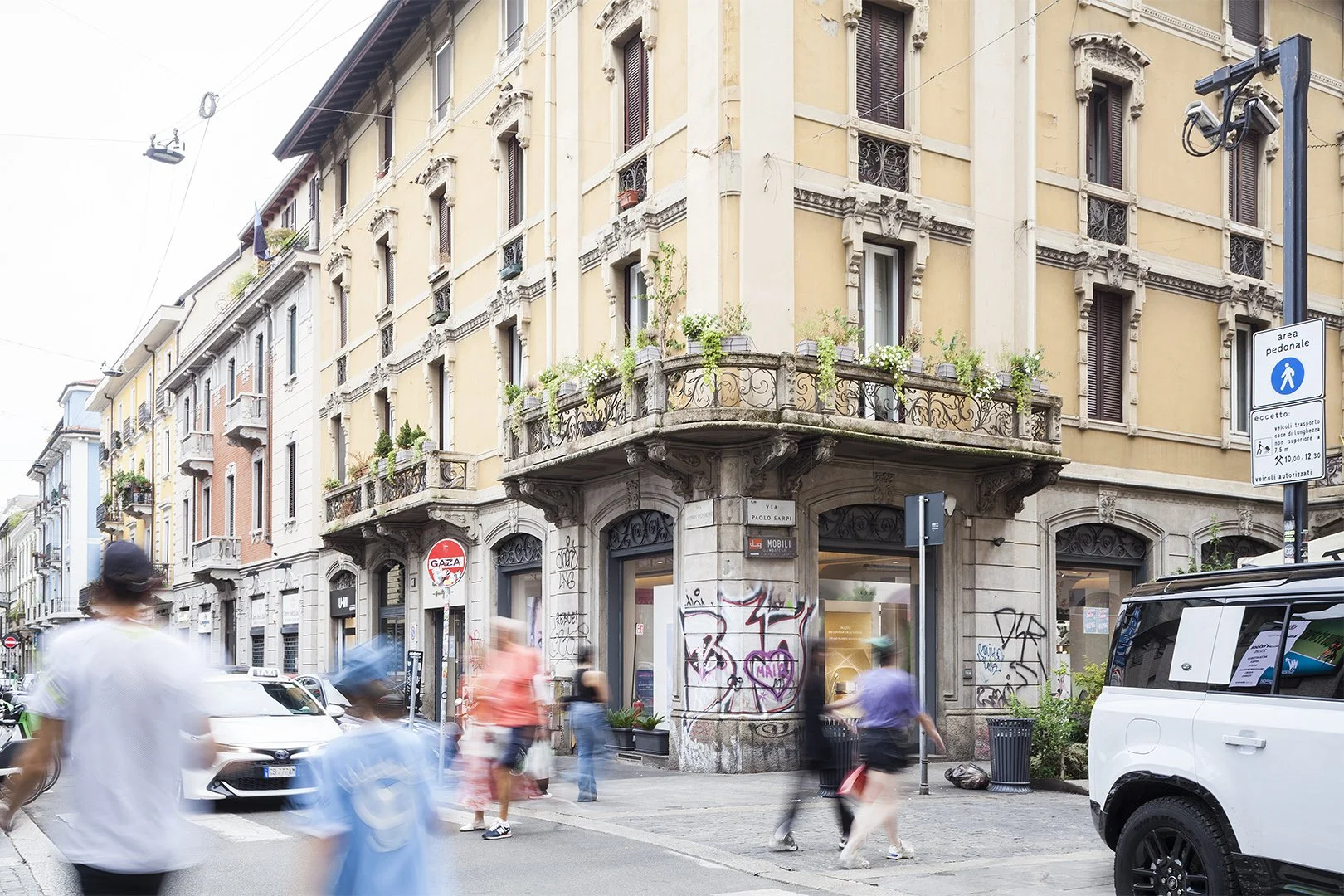 Edificio storico con balcone fiorito e negozio al piano terra, strada trafficata con persone e veicoli in movimento, cartelli stradali e pubblicitari.