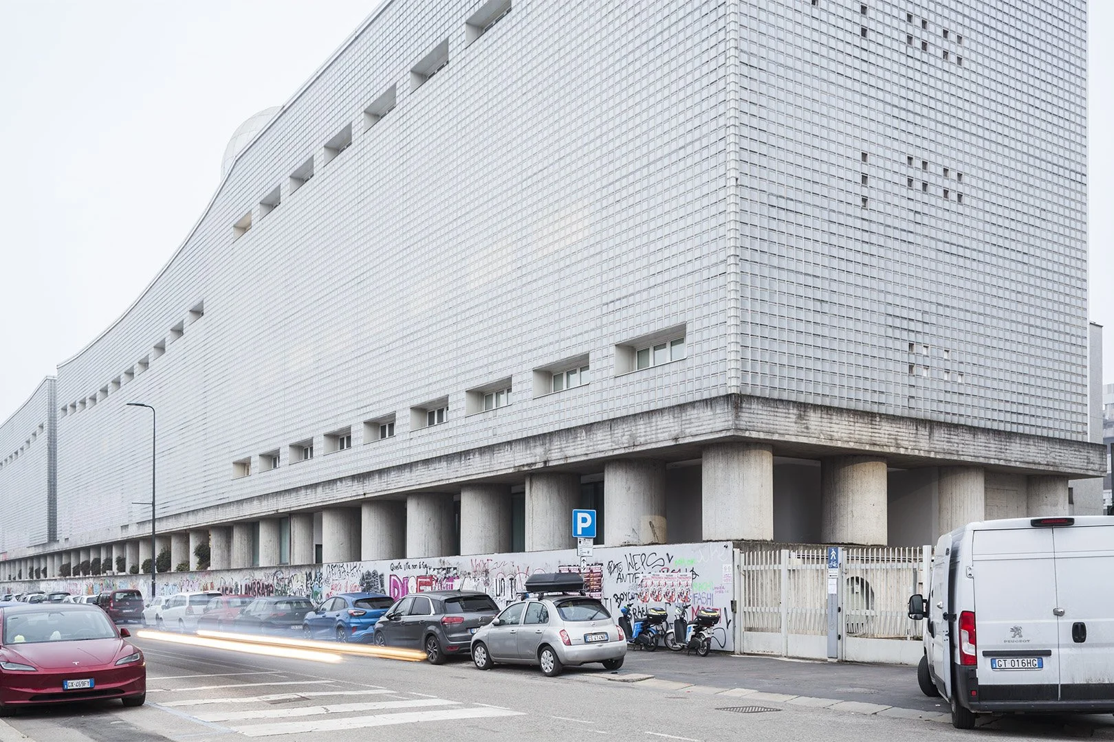 Edificio moderno con facciata di mattonelle bianche, colonne di cemento e graffiti sulla barriera di delimitazione del parcheggio. Auto parcheggiate lungo la strada. Strada urbana con strisce pedonali.