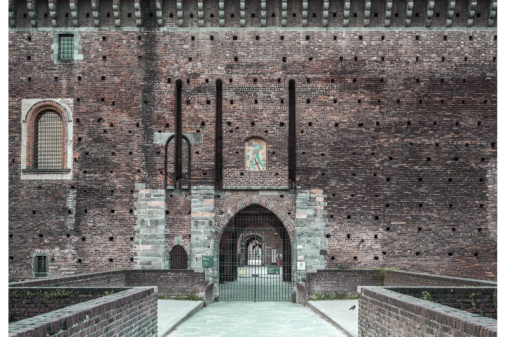 Una grande mura di mattoni rossi con una porta d'ingresso in metallo nero e un cancello che la protegge. Sulla parete ci sono finestre con inferriate e un'illustrazione di un drago.