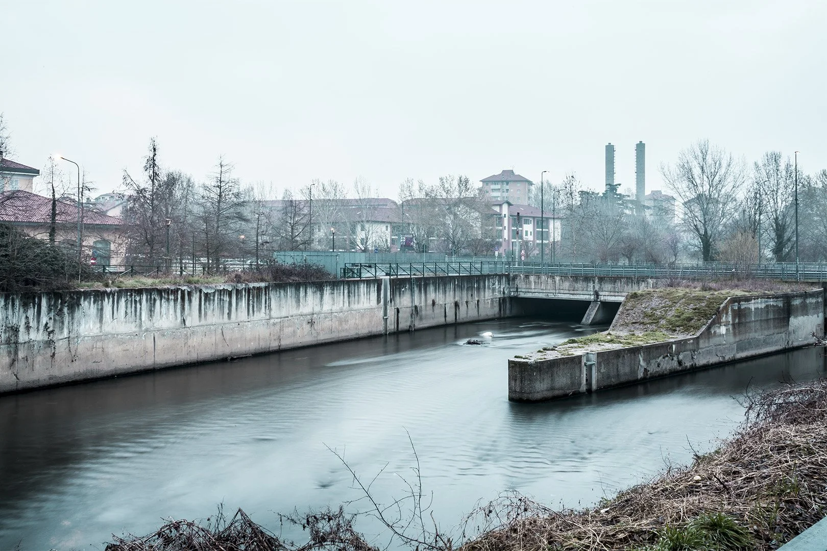 Fiume urbano con acque calme, sponde in cemento e edifici residenziali sullo sfondo, atmosfera fosca e brumosa.