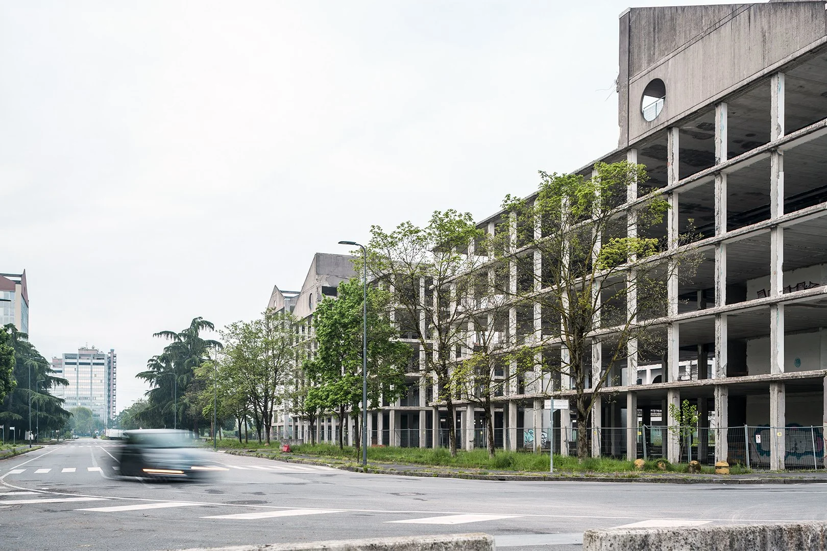 Edificio incompleto lungo una strada con alberi e un'auto in movimento.