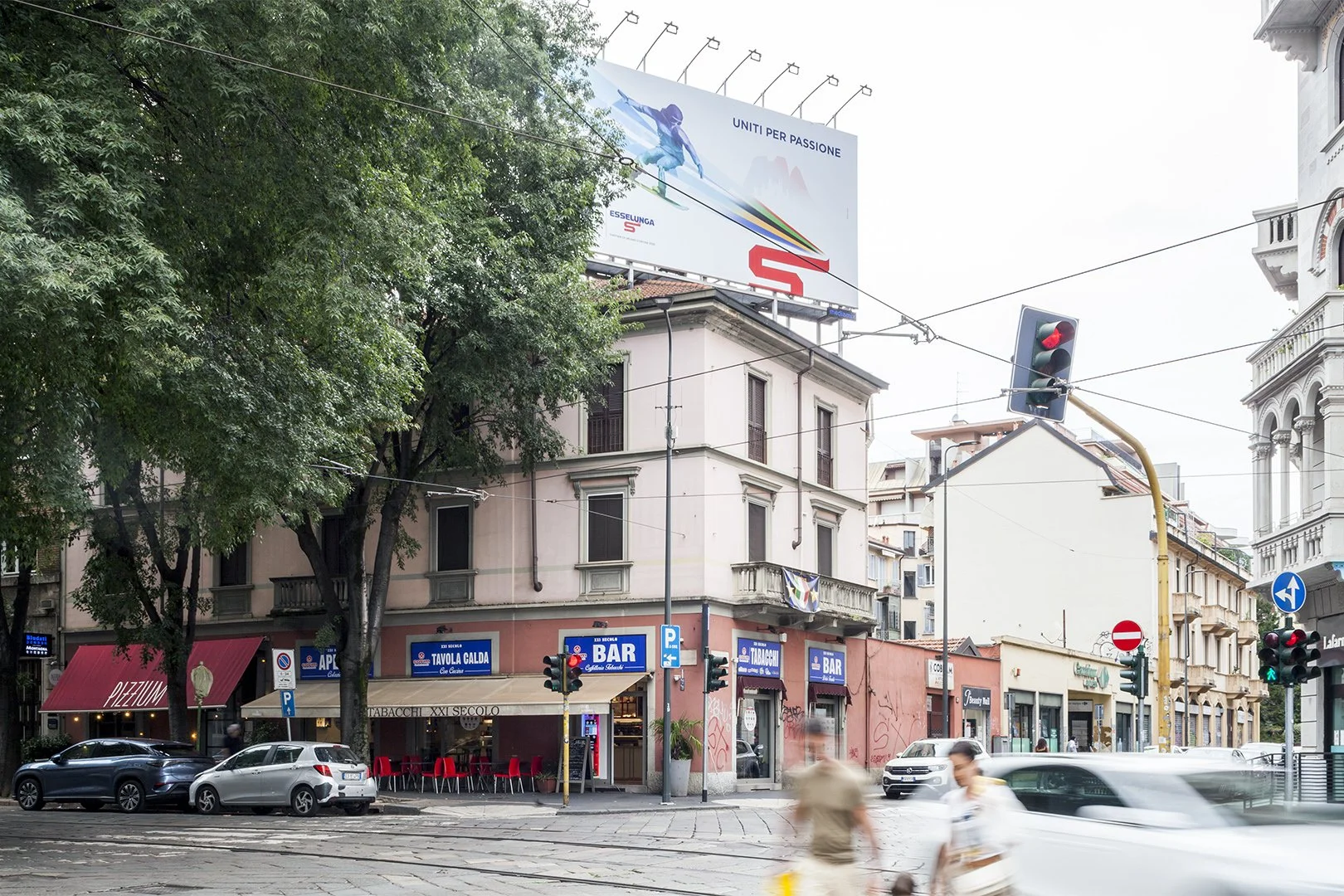 Una strada cittadina con edifici, negozi, automobili e passanti. Sorprendente cartellone pubblicitario con una persona arrampicata e una scritta "Unità per Passione". La scena è cittadina, con traffico e luci di samba.