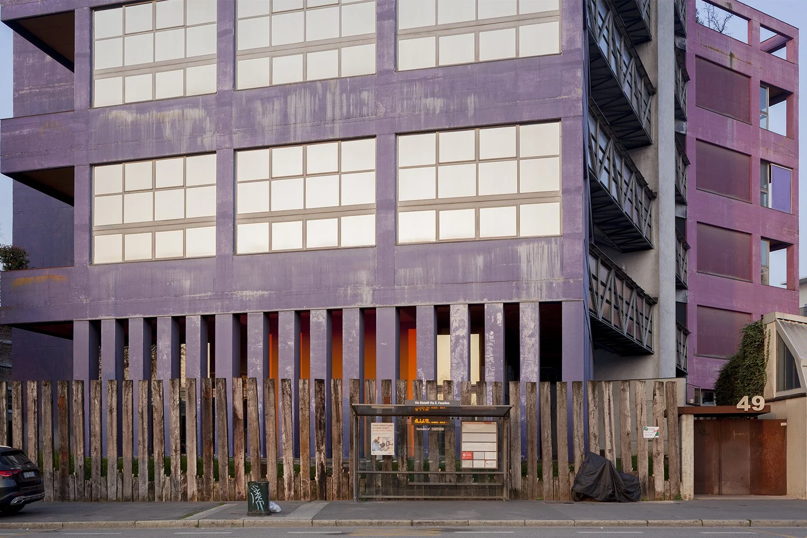 Edificio moderno con struttura in cemento di colore viola, grandi finestre e una scala esterna in metallo nero sul lato destro. In primo piano, fermata dell'autobus con scheda informativa, una recinzione di assi di legno e un'auto parcheggiata sulla 