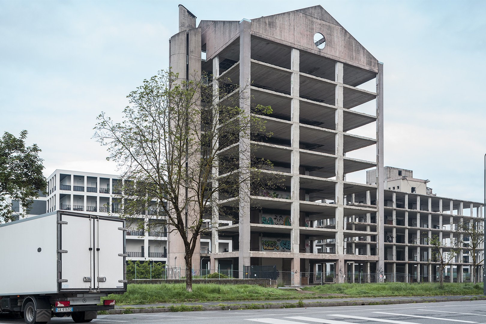 Edificio abbandonato in costruzione con strutture di cemento e graffiti sulle pareti, fianco strada con parcheggio e camion