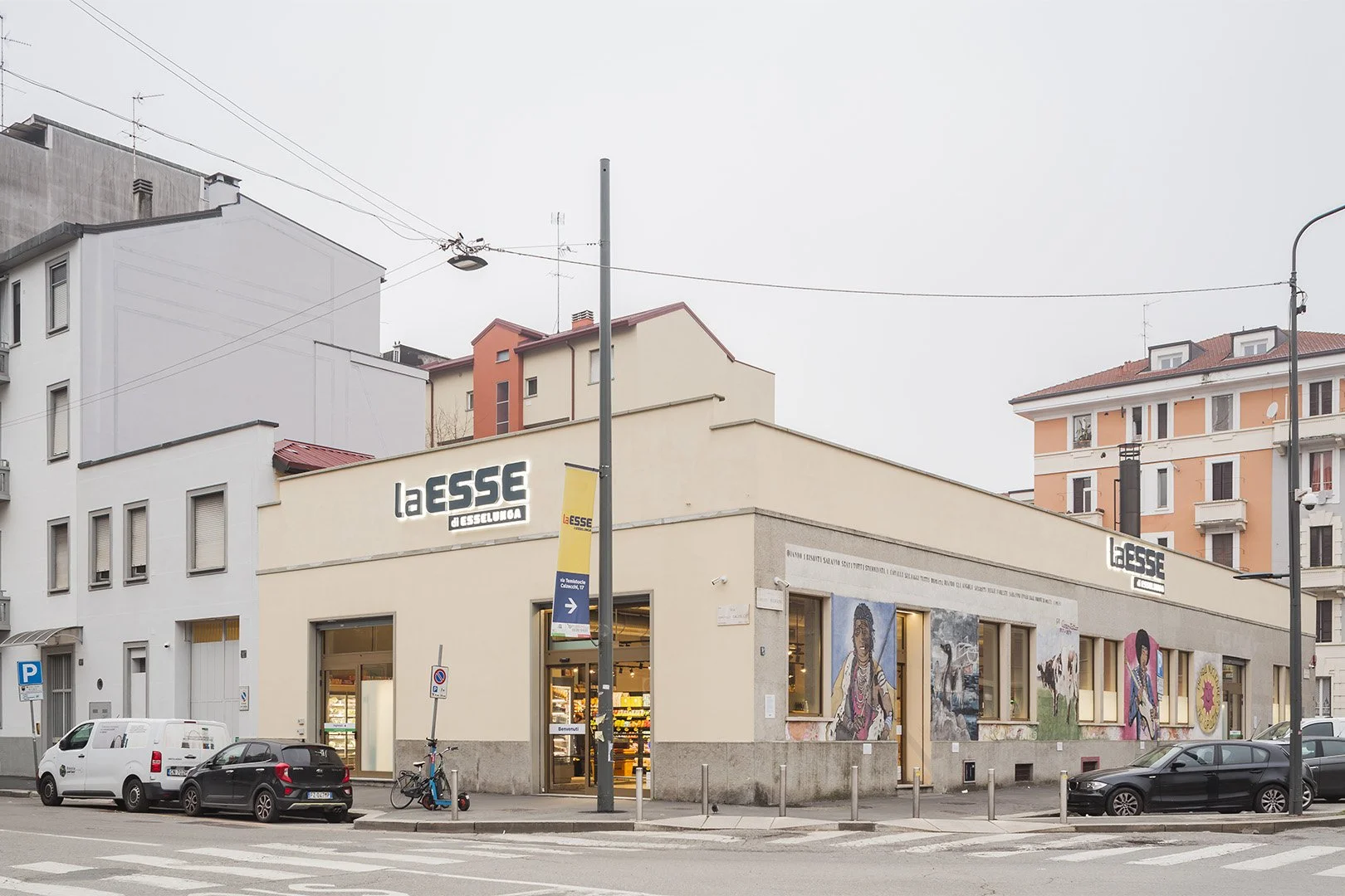 Esterno di un supermercato chiamato La Esse a Desselunga, con muri decorati con murales e diverse auto parcheggiate di fronte