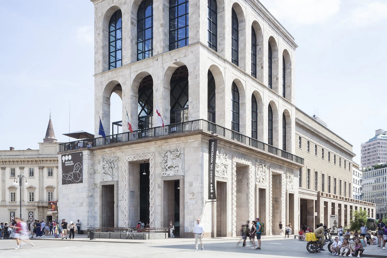 Edificio storico in stile neoclassico con archi e bassorilievi, sede del Museo del Novecento, in Piazza del Duomo a Milano, con persone che camminano e si fermano nell'area circostante