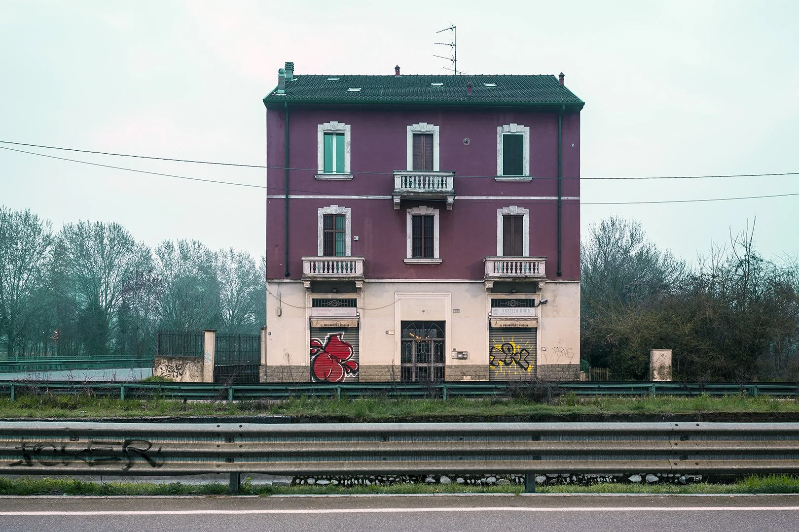 Edificio di tre piani di colore rosso con balconi, subito sopra una strada, con graffiti sul muro al piano terra, e alberi sullo sfondo.