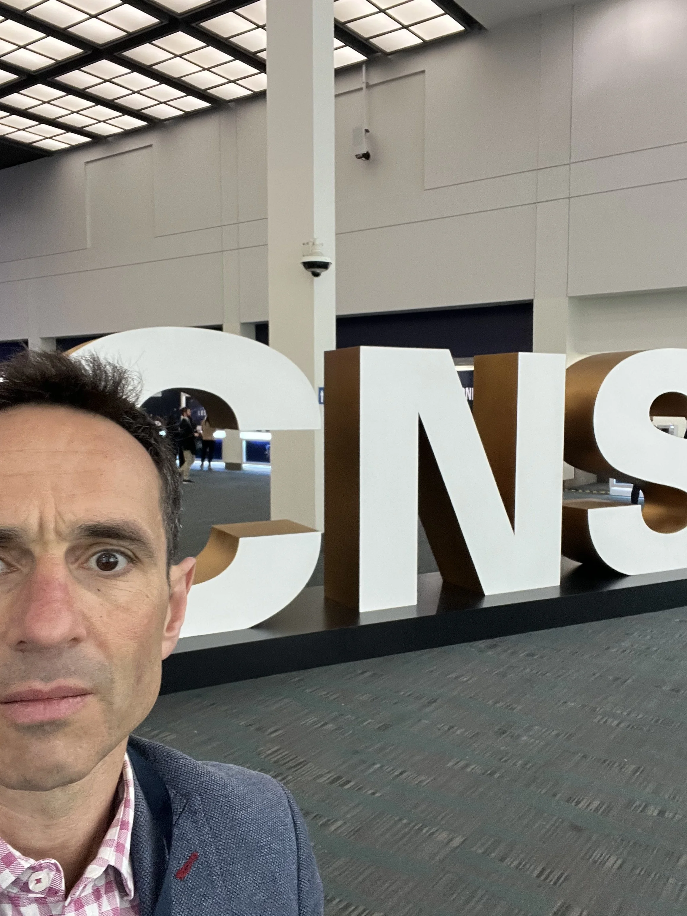 A man taking a selfie at an airport terminal in front of large white and gold letters spelling 'CES'.