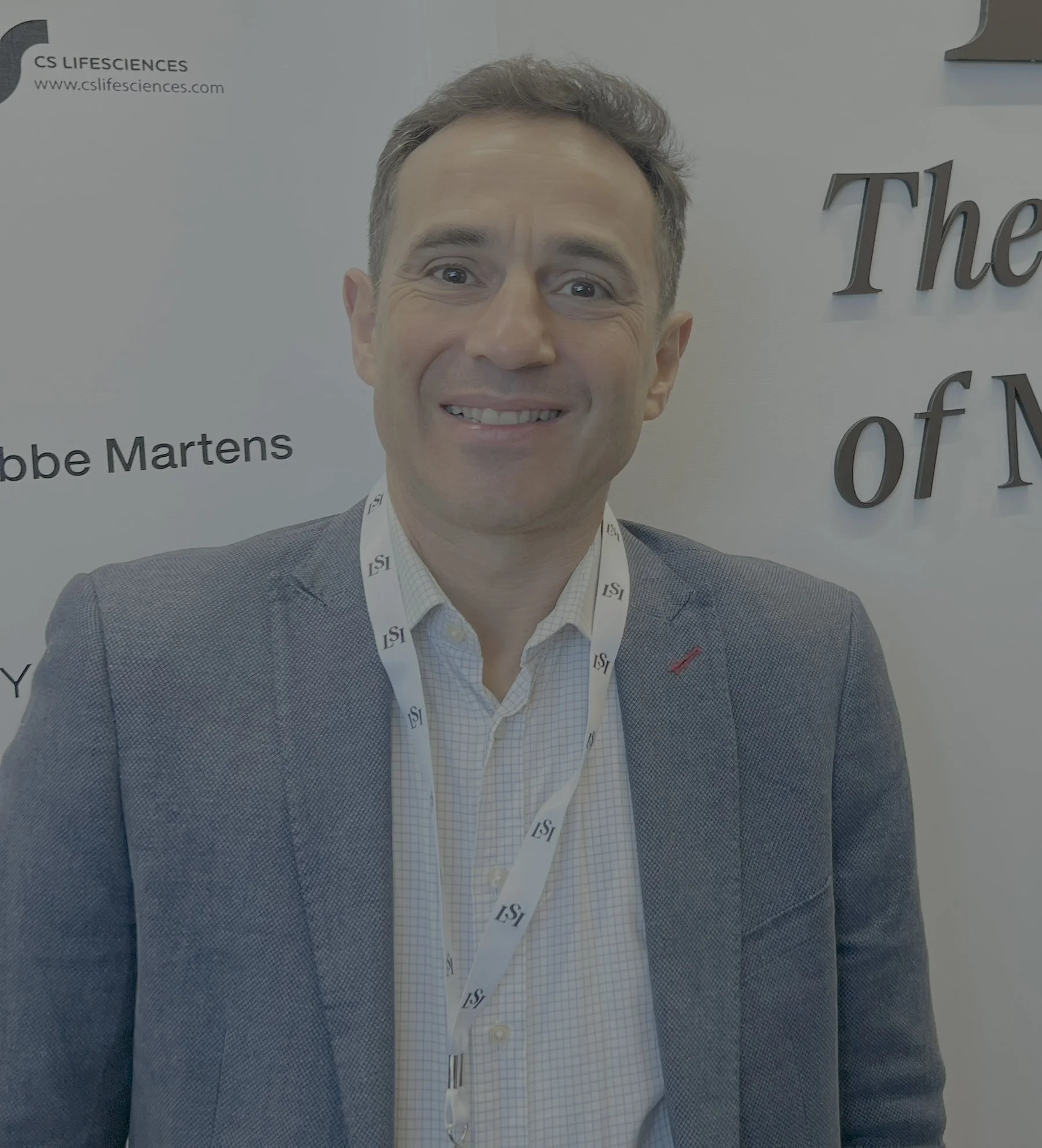 A smiling man in a gray blazer and white checkered shirt, wearing a lanyard with 'LSI' on it, standing in front of a white background with text about CS Lifesciences and an event titled 'The of N'