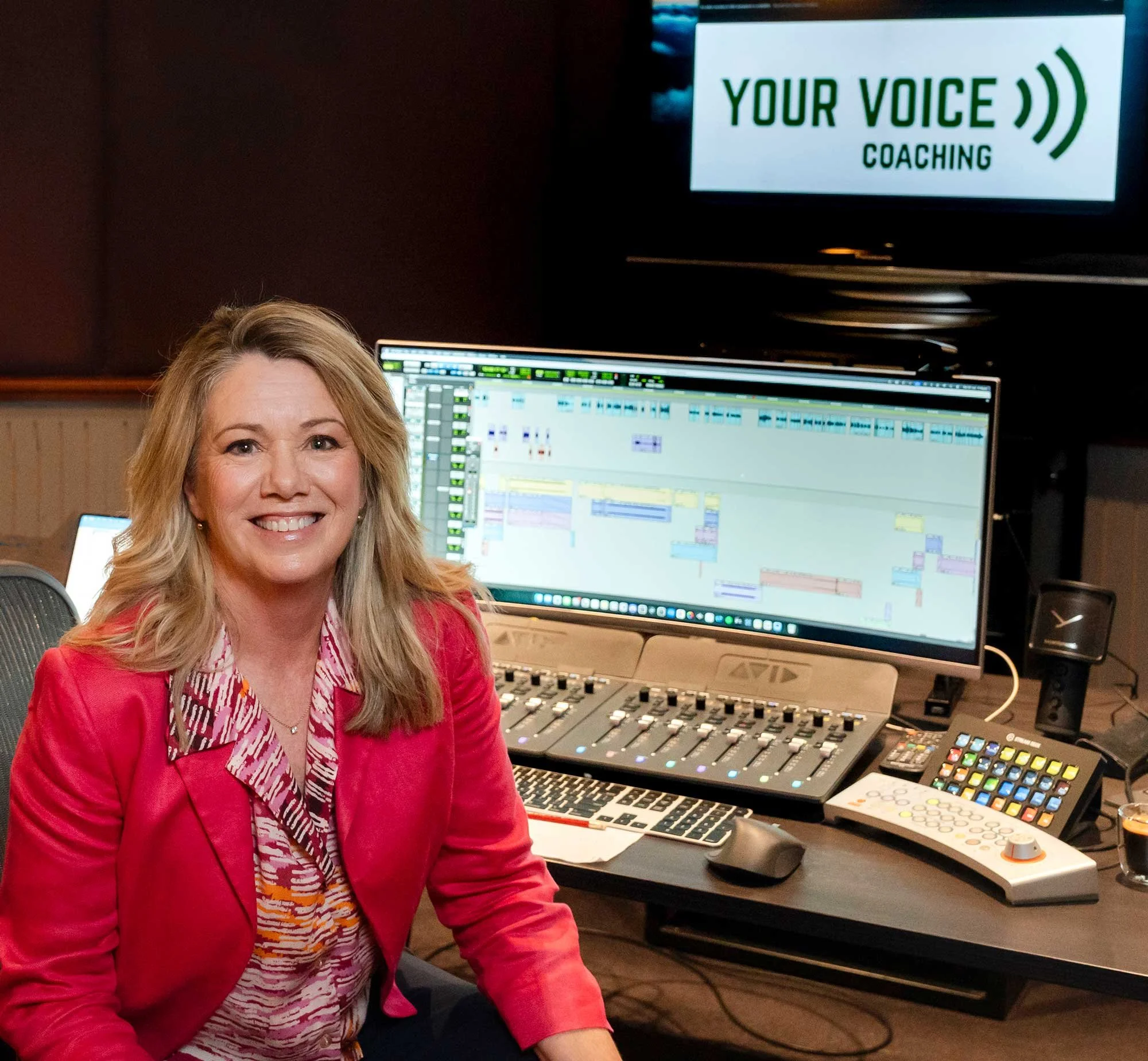 A woman with blonde hair and a red blazer smiling in a radio or recording studio with computer monitors and audio equipment, and a sign in the background that reads 'Your Voice Coaching'.