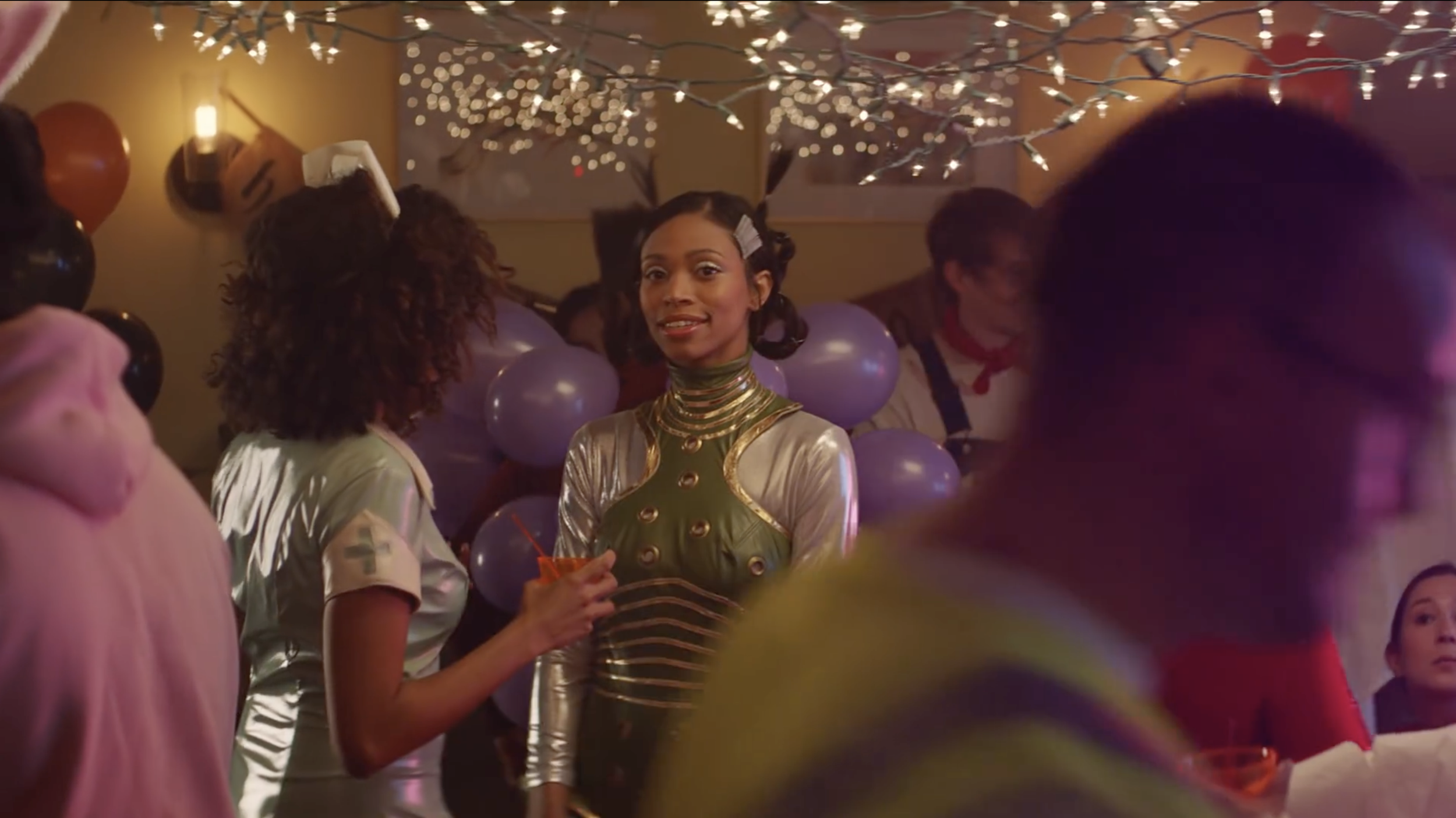 A woman with dark hair styled in vintage curls, wearing a shiny green and gold futuristic costume, holds a drink at a party with purple balloons and string lights.