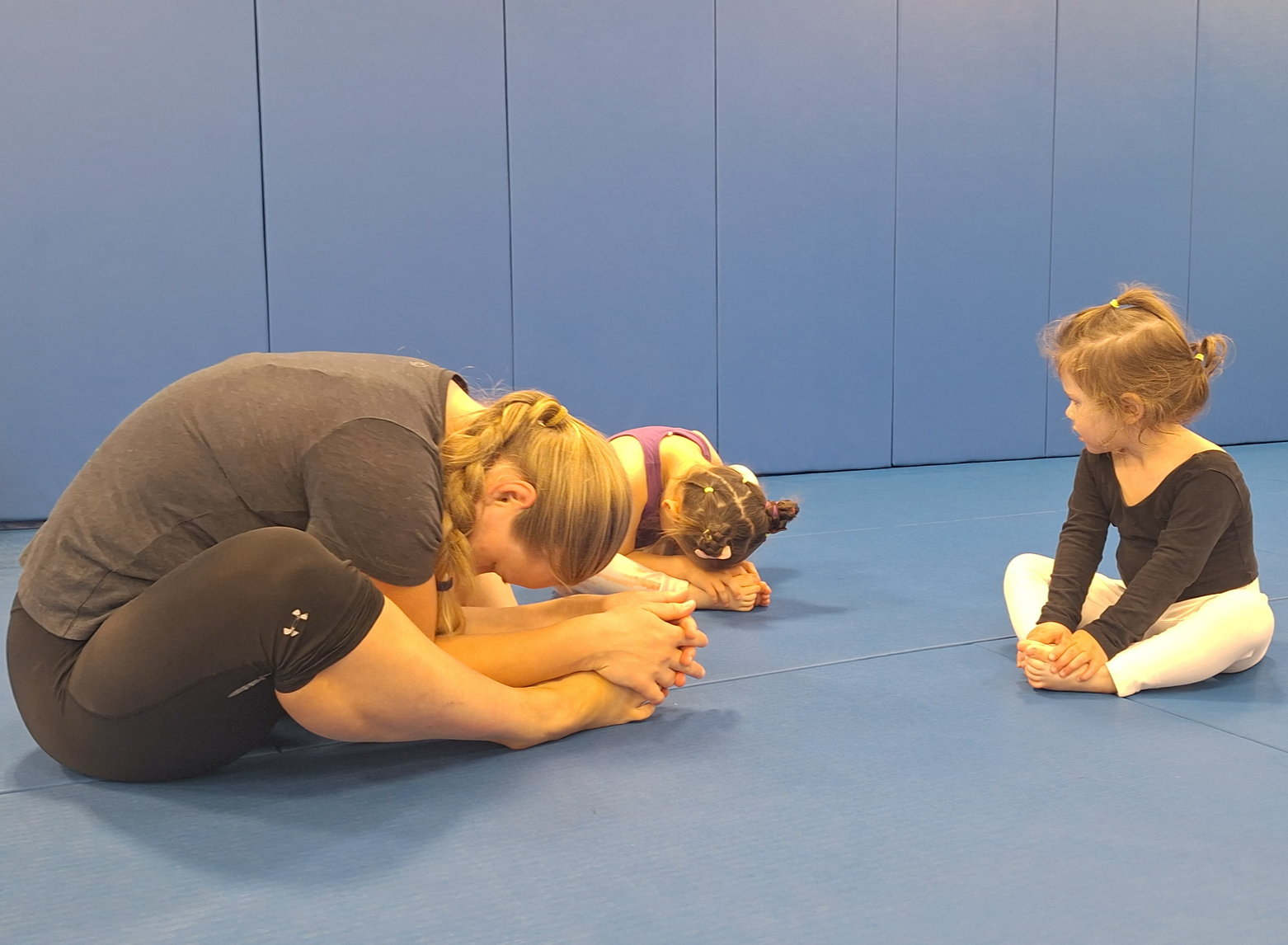 Two adults and two children practicing seated forward bend stretches on a blue gym mat in a gym or martial arts studio, with a blue padded wall in the background.