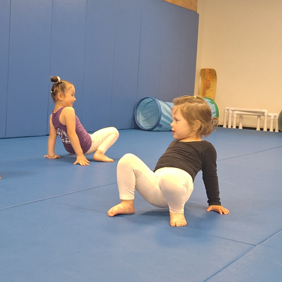 Two young girls in athletic clothing playing and climbing on a blue padded gym floor, with a blue padded wall and play tunnel in the background.