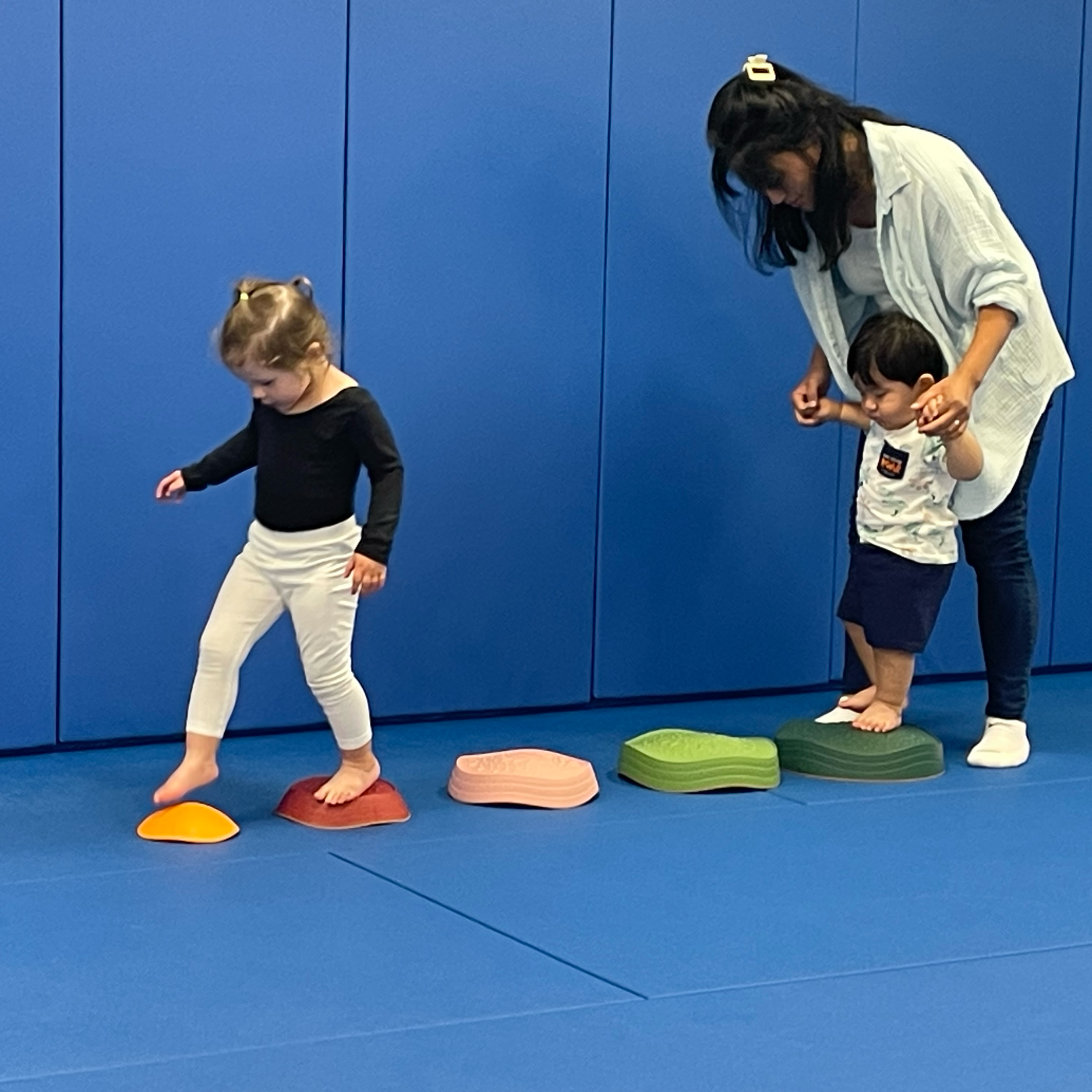 A woman and two children practicing balance on colored stepping stones in a room with blue padded walls.