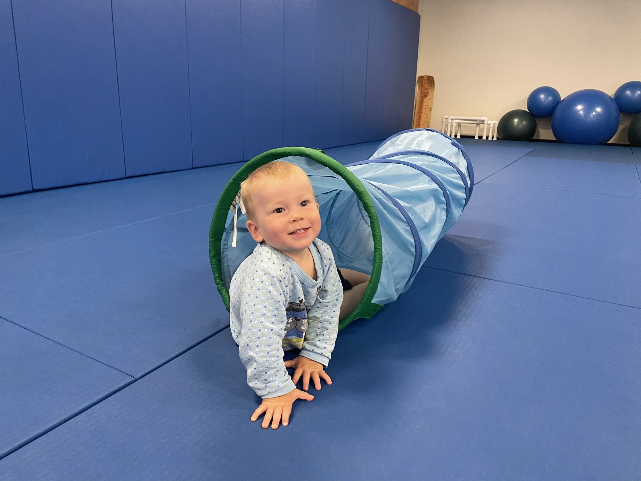 A young child with blonde hair smiling while crawling on blue padded gym floor with a blue play tunnel and exercise balls in the background.
